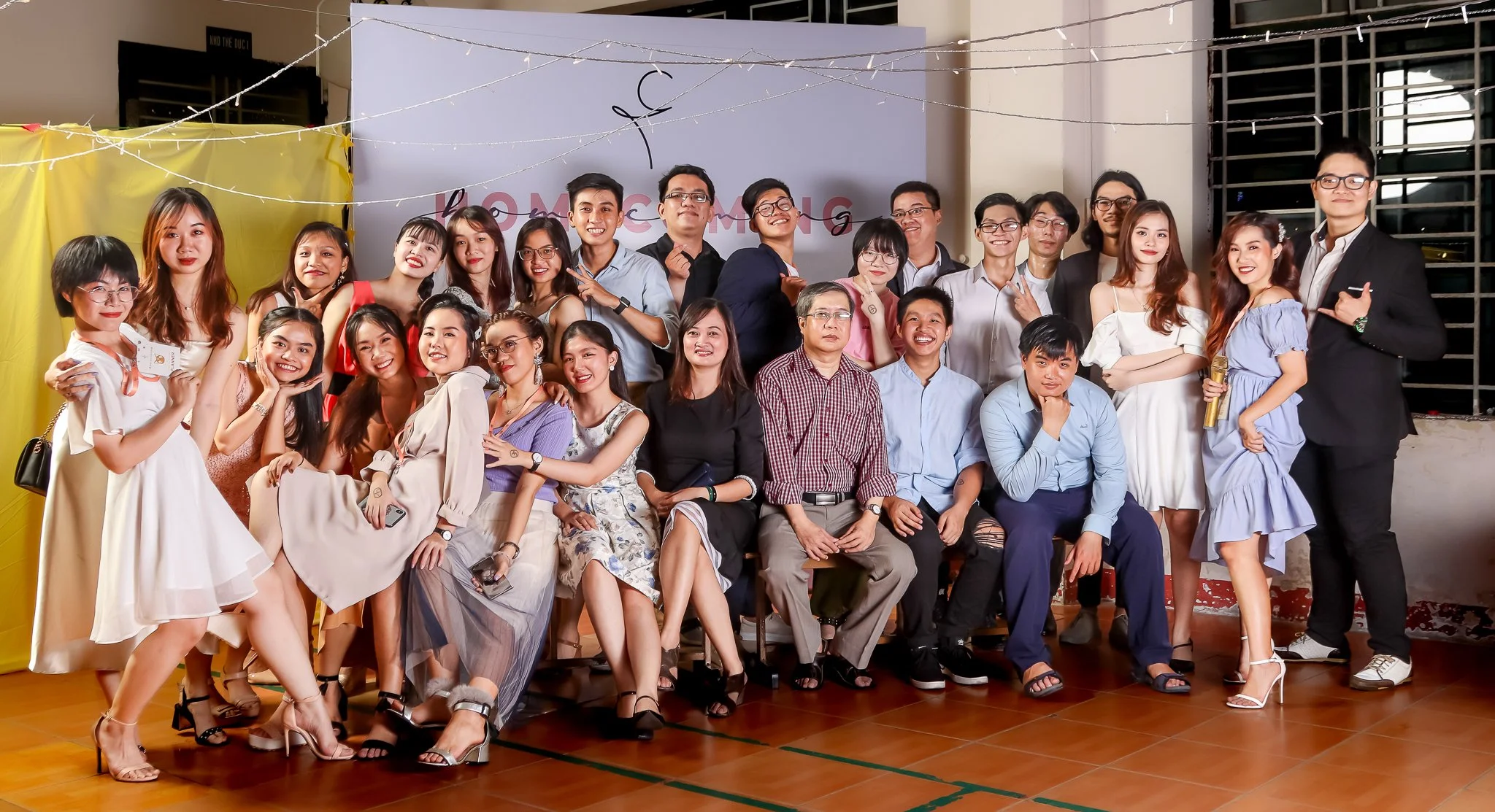 Group of people celebrating at a formal event, with smiling faces and various stylish outfits, inside a decorated hall with a yellow curtain and string lights.