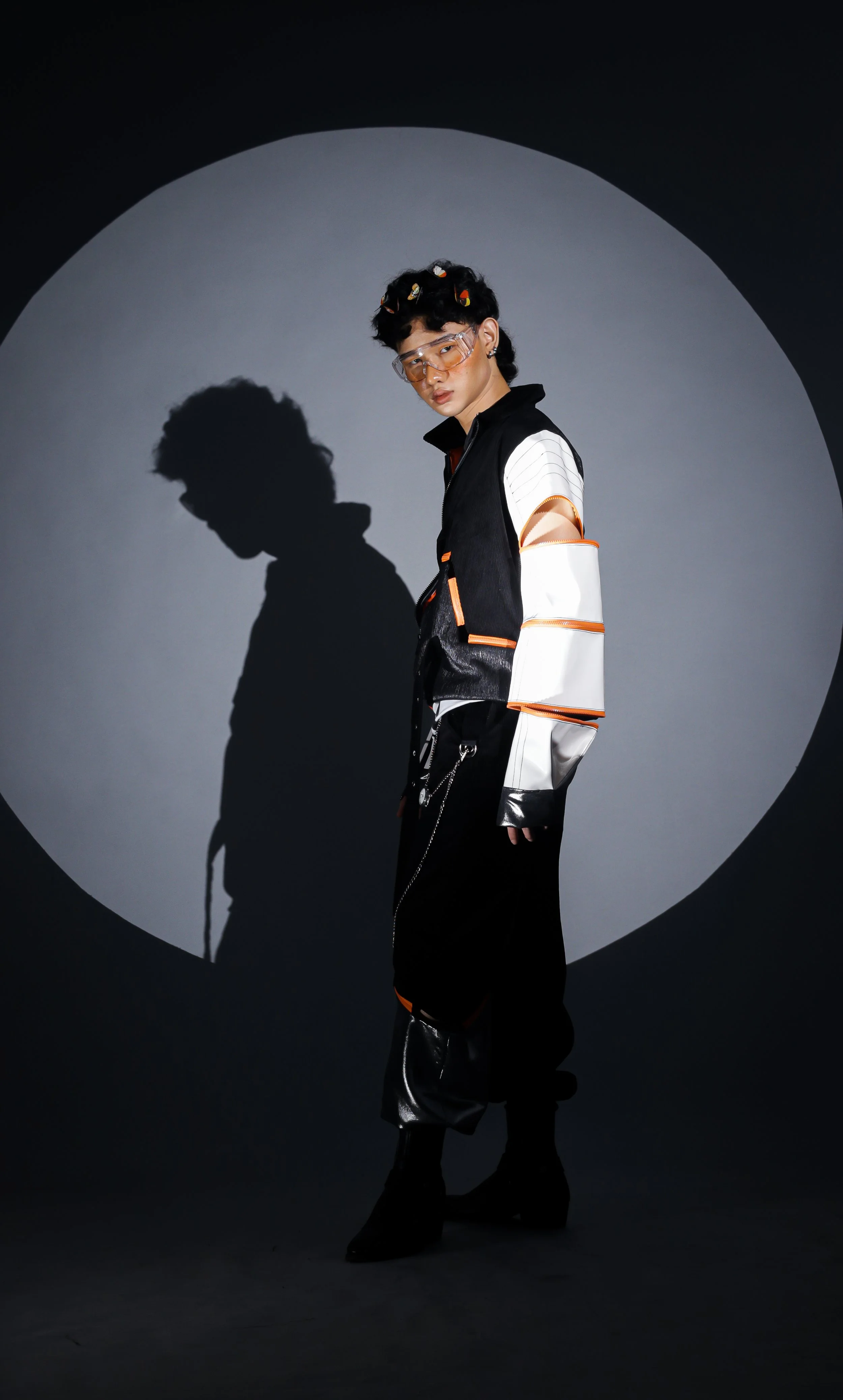 A young woman with short curly hair and colorful hair clips, wearing futuristic glasses, stands in front of a dark background with a spotlight creating a shadow. She is dressed in a stylish, modern black and white outfit with metallic accents.