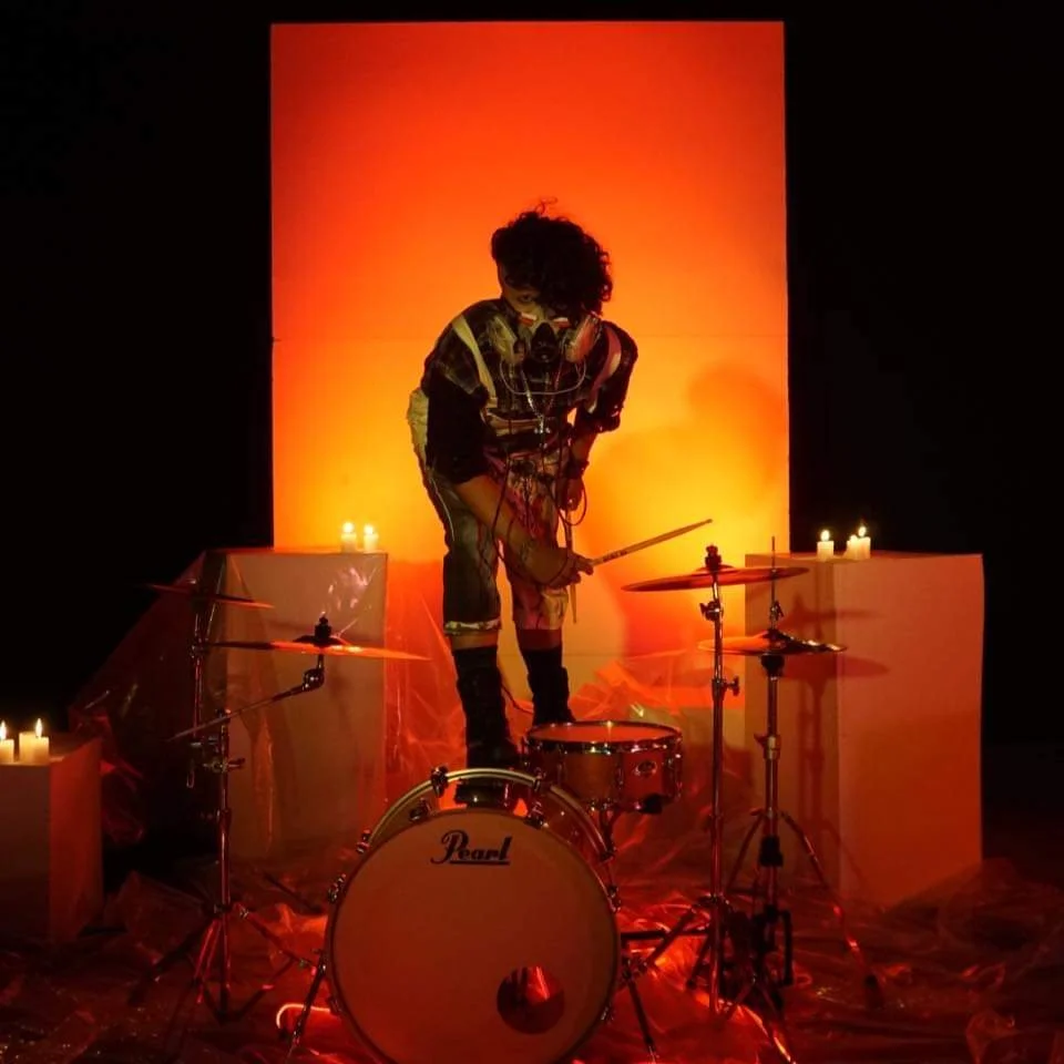 Person with curly hair playing drums on a stage illuminated with orange lighting, surrounded by candles.