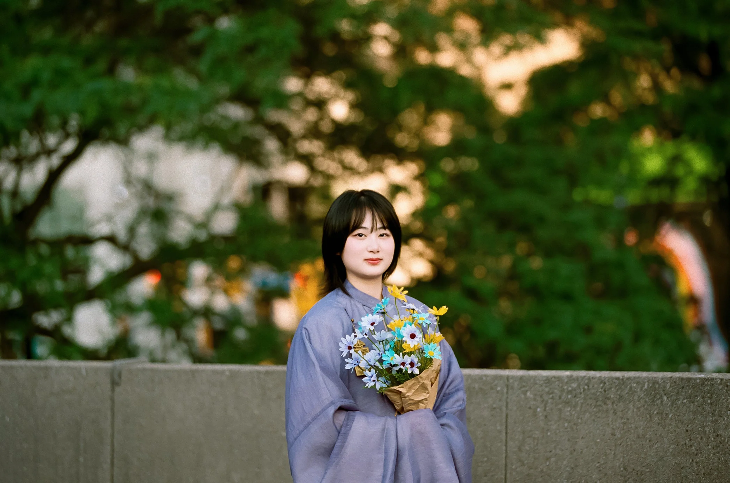 Young woman in a purple coat holding a bouquet of colorful artificial flowers, standing outdoors against a background of green trees and soft sunset light.