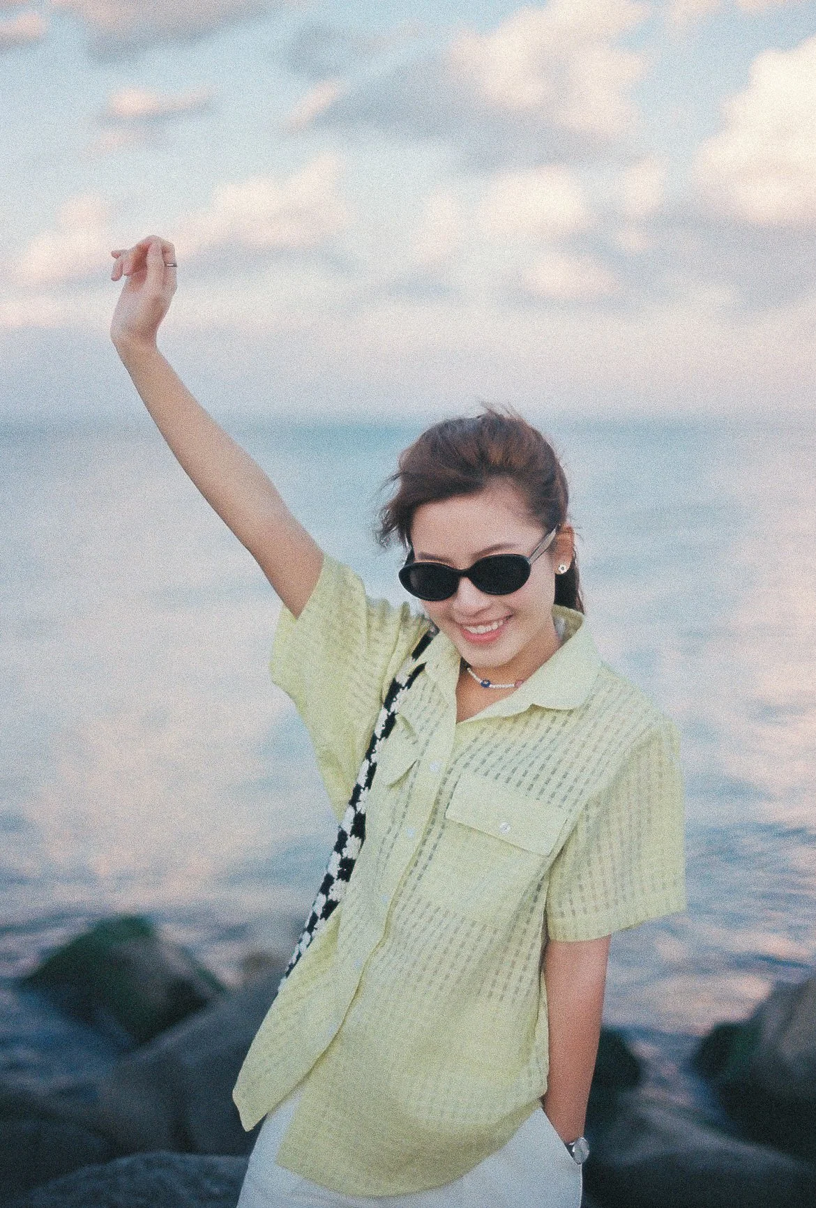 A woman wearing black sunglasses, a light green shirt, and a black-and-white patterned bag posing outdoors near water with rocks and a cloudy sky.