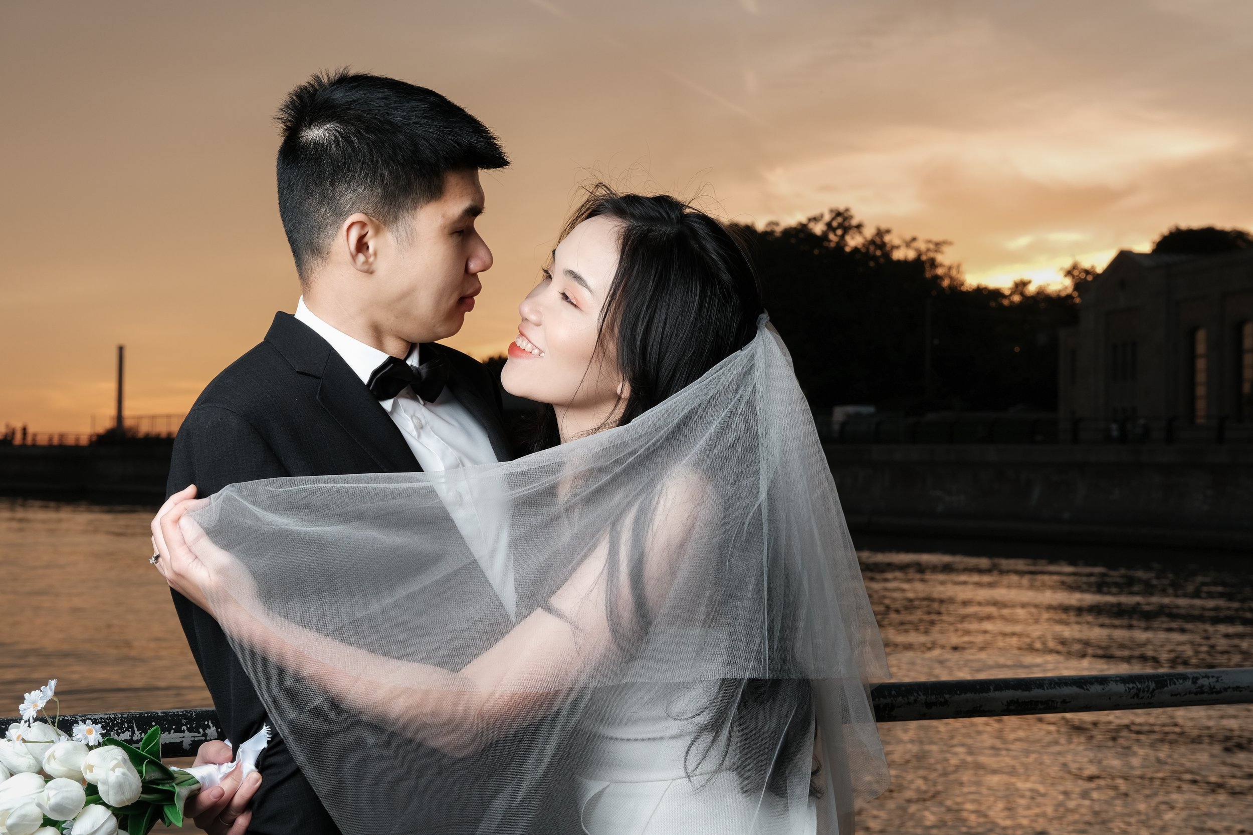 Couple on their wedding day, standing near a body of water at sunset. The bride wears a white wedding gown and veil, and the groom is dressed in a black tuxedo. The bride holds a bouquet of white flowers.