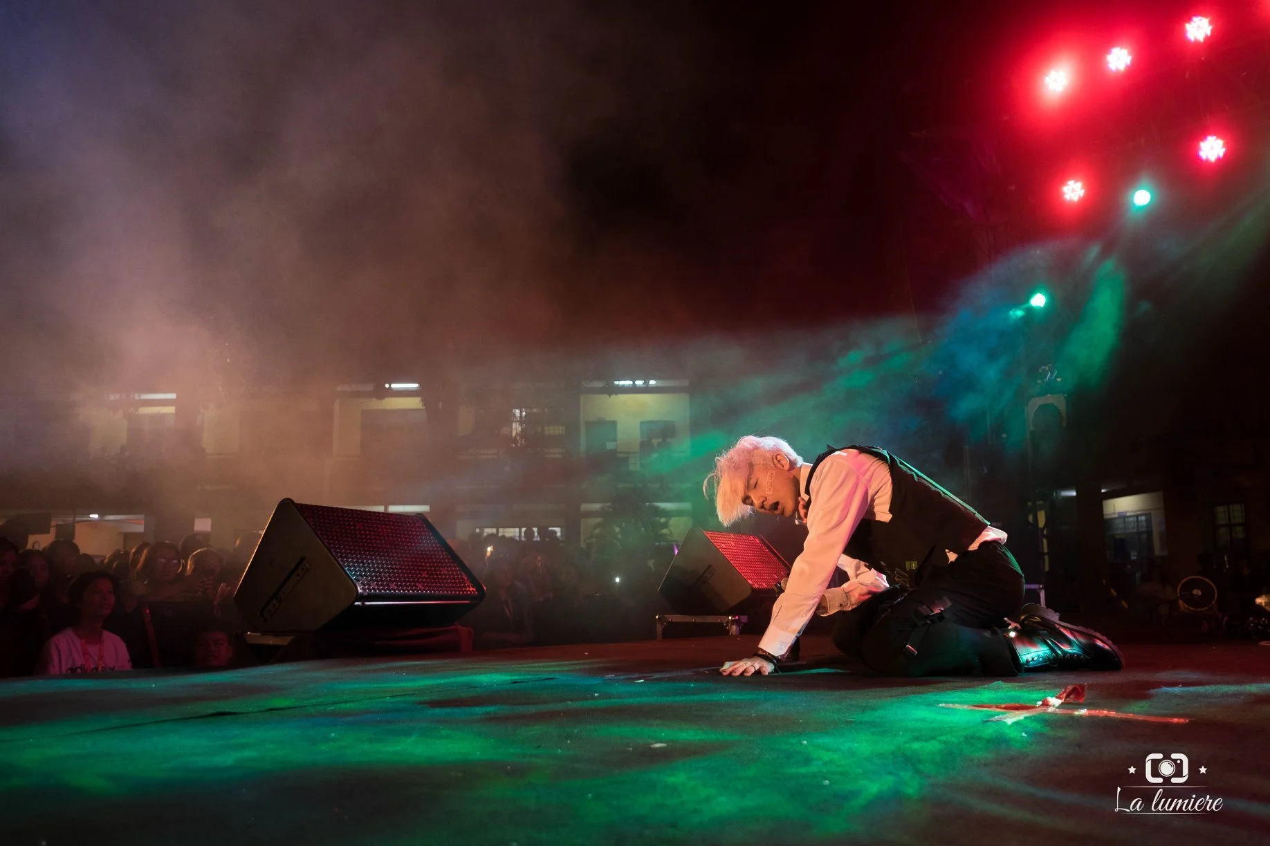 Performer kneeling on stage with one hand on the ground, appearing emotional during a concert with colorful stage lighting and an audience in the background.