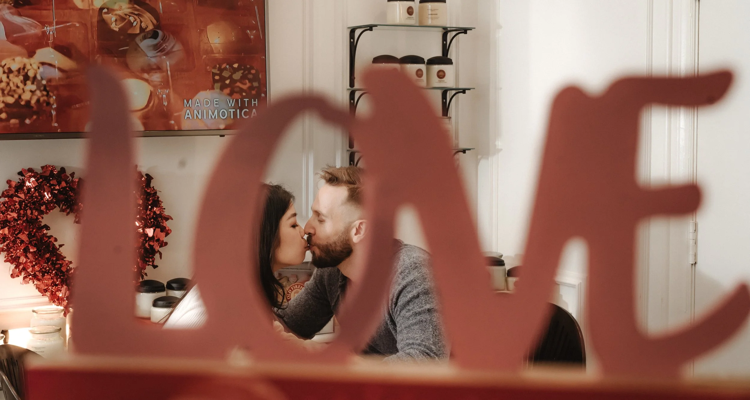 A couple kissing indoors, seen through a decorative red word 'LOVE' cutout in the foreground. The background includes shelving with jars and a Valentine's Day heart decoration.