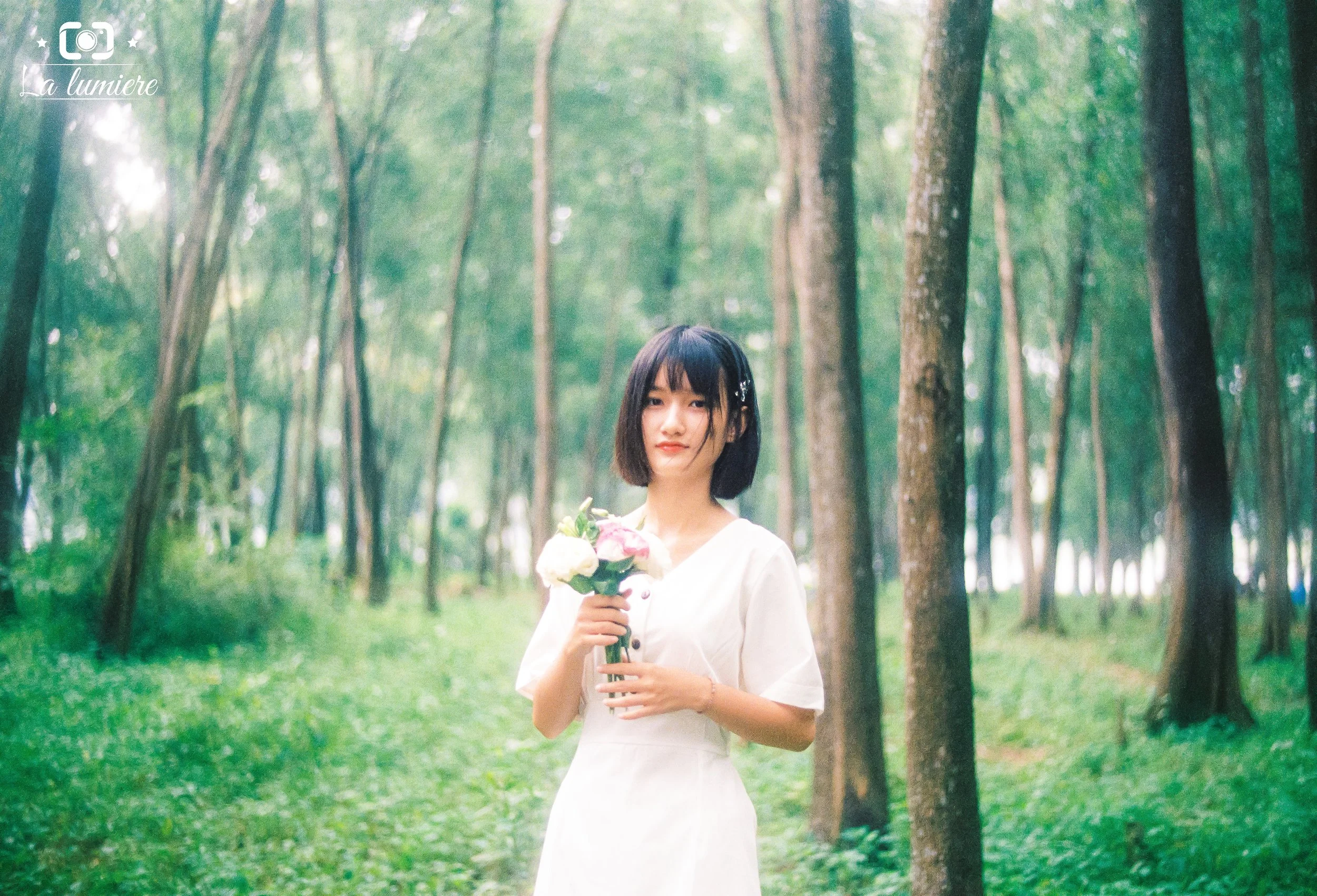 A young woman with short black hair holding a bouquet of flowers in a lush green forest.