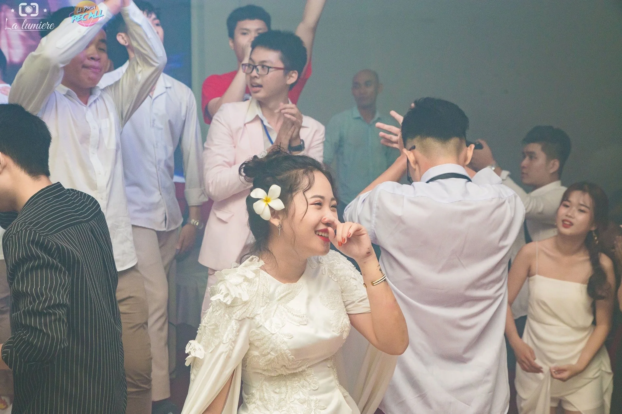 People dancing and celebrating at a party or wedding reception, with a woman in a white dress and flower in her hair smiling and a man with glasses in front of her, along with other guests in the background.
