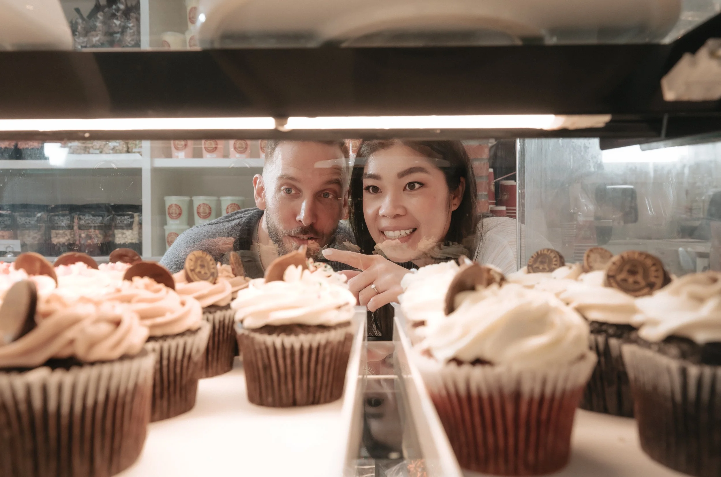 Two people looking at cupcakes through a glass display, smiling and pointing at the desserts.