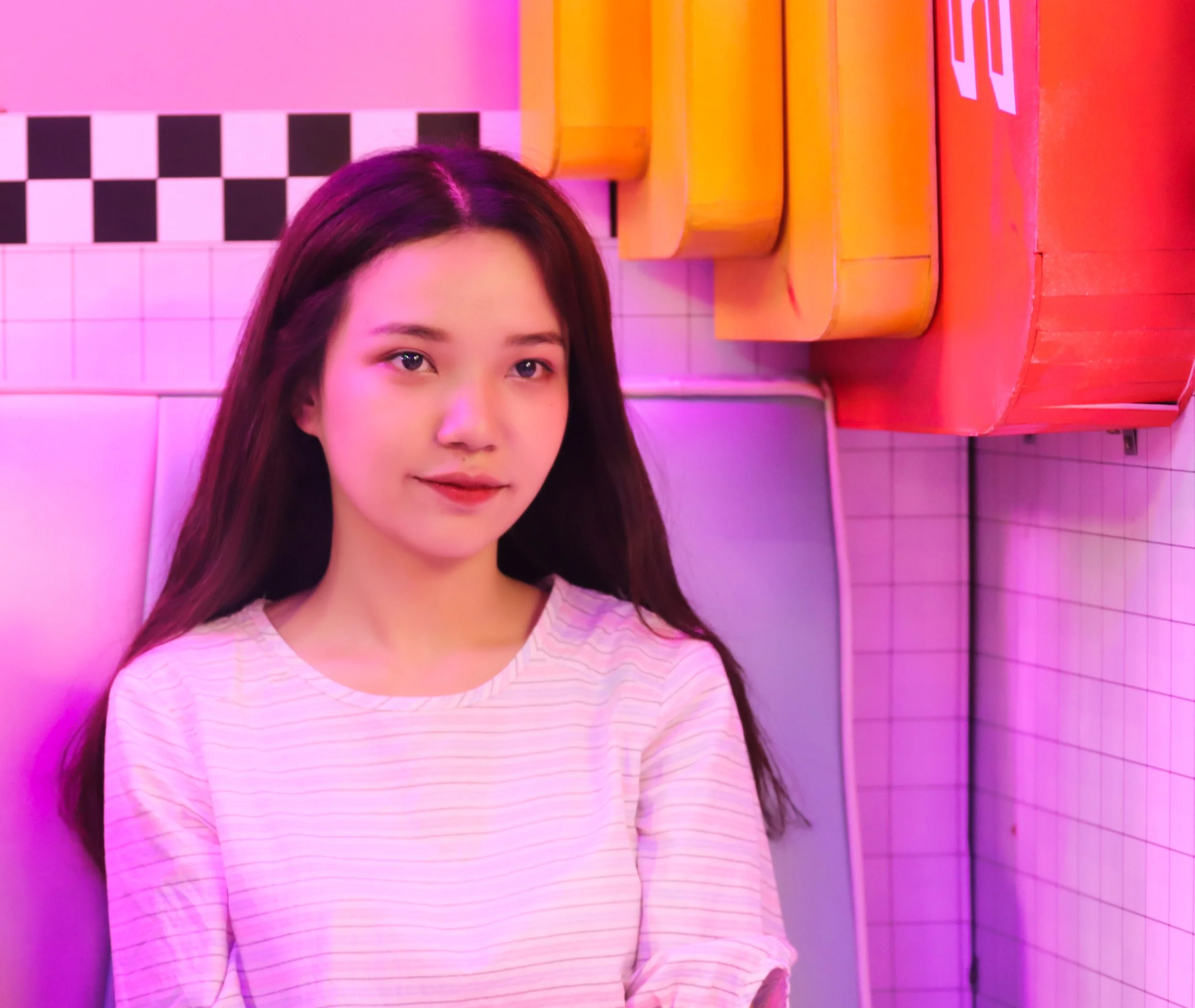 A young woman with long dark hair wearing a light pink striped shirt sitting against pink-tiled wall with colorful signs above in a neon-lit setting.