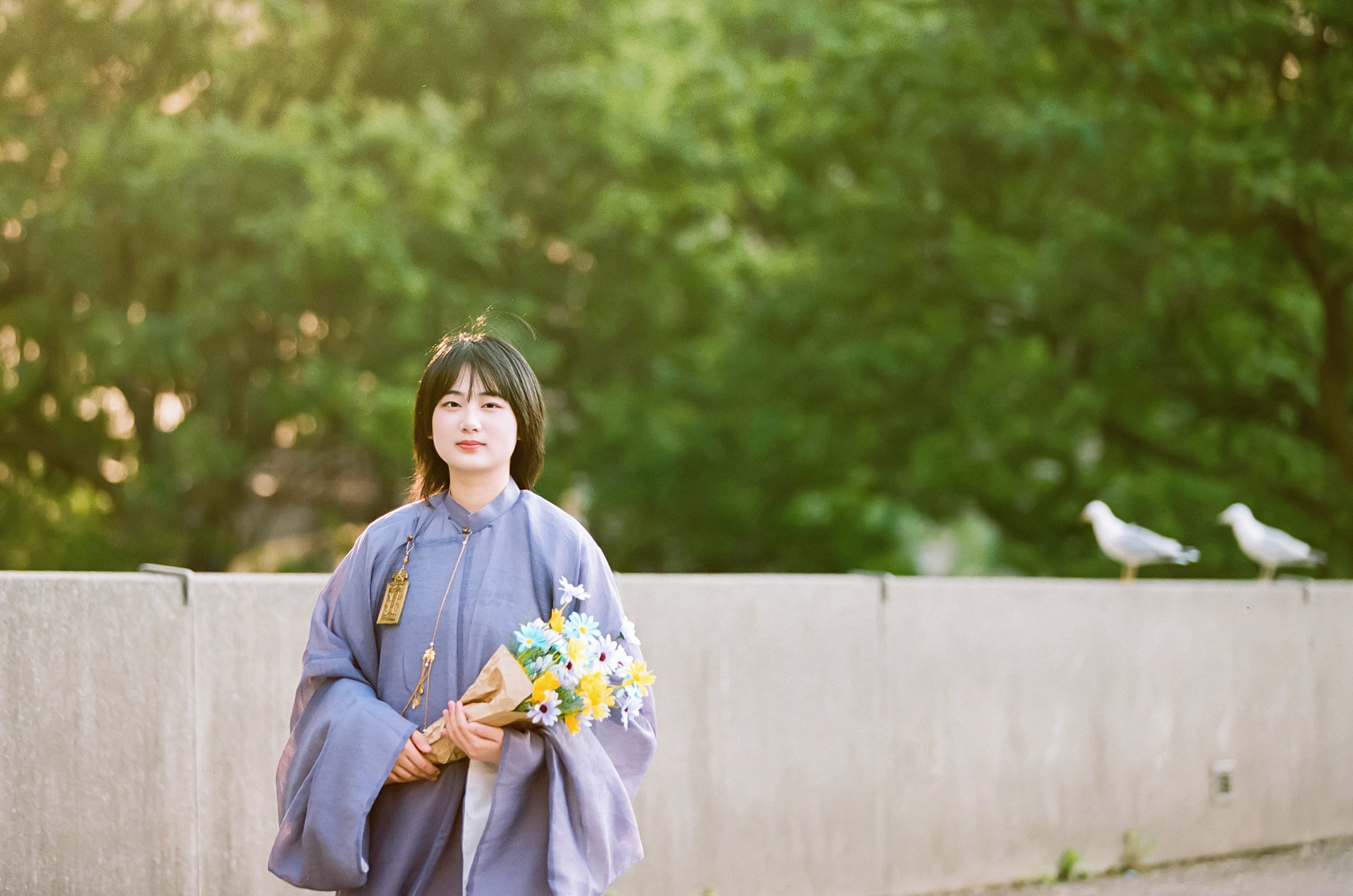 A young woman in a light gray graduation gown holding a bouquet of flowers, standing outdoors in front of a concrete wall with green trees and two white birds in the background.