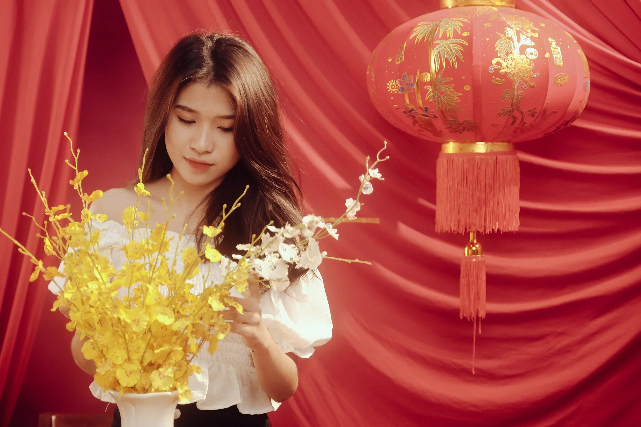 A young woman with long dark hair holding a bouquet of yellow and white flowers, standing in front of a red curtain with a large red Chinese lantern hanging nearby.