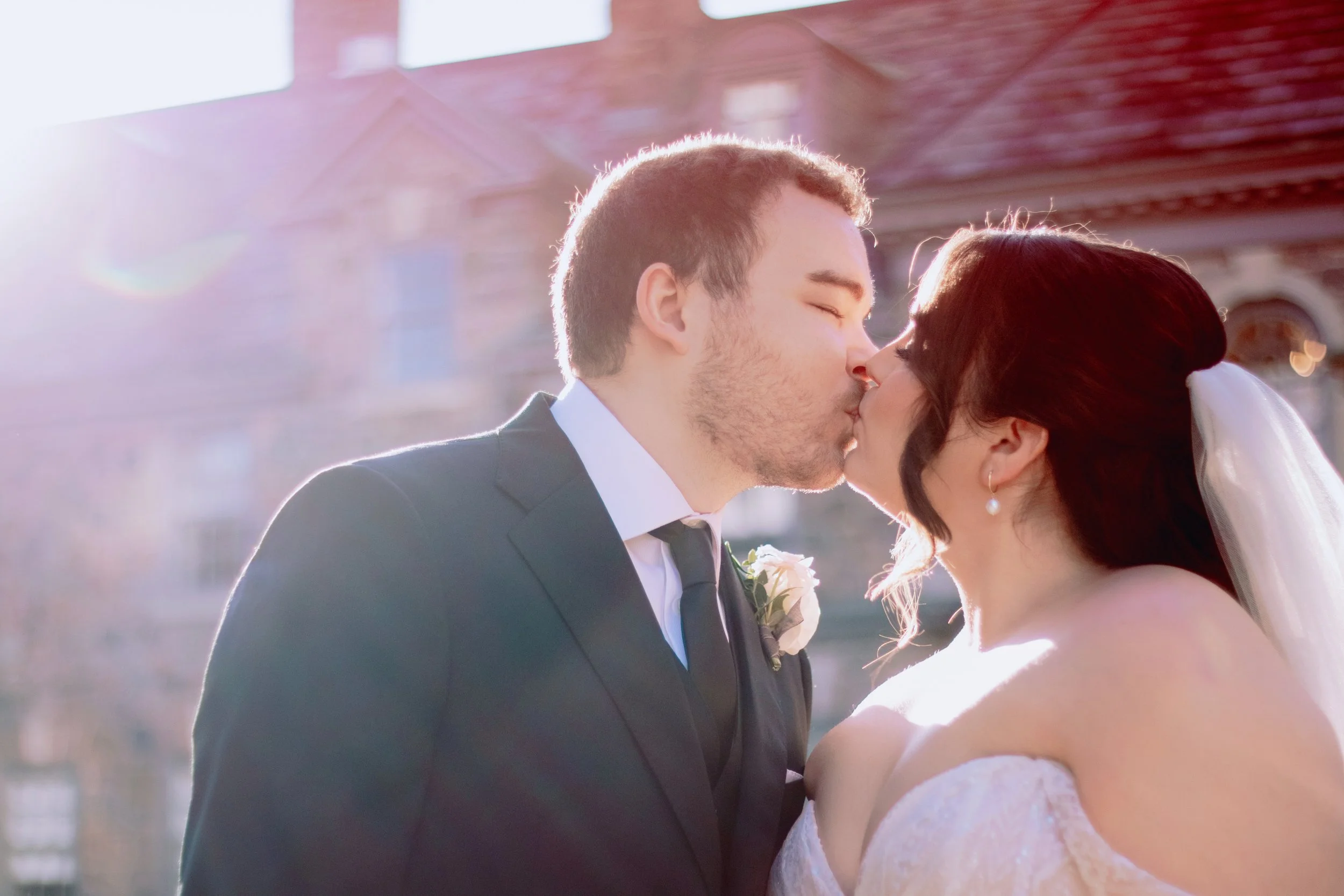 A bride and groom kissing outdoors with sunlight shining in the background.