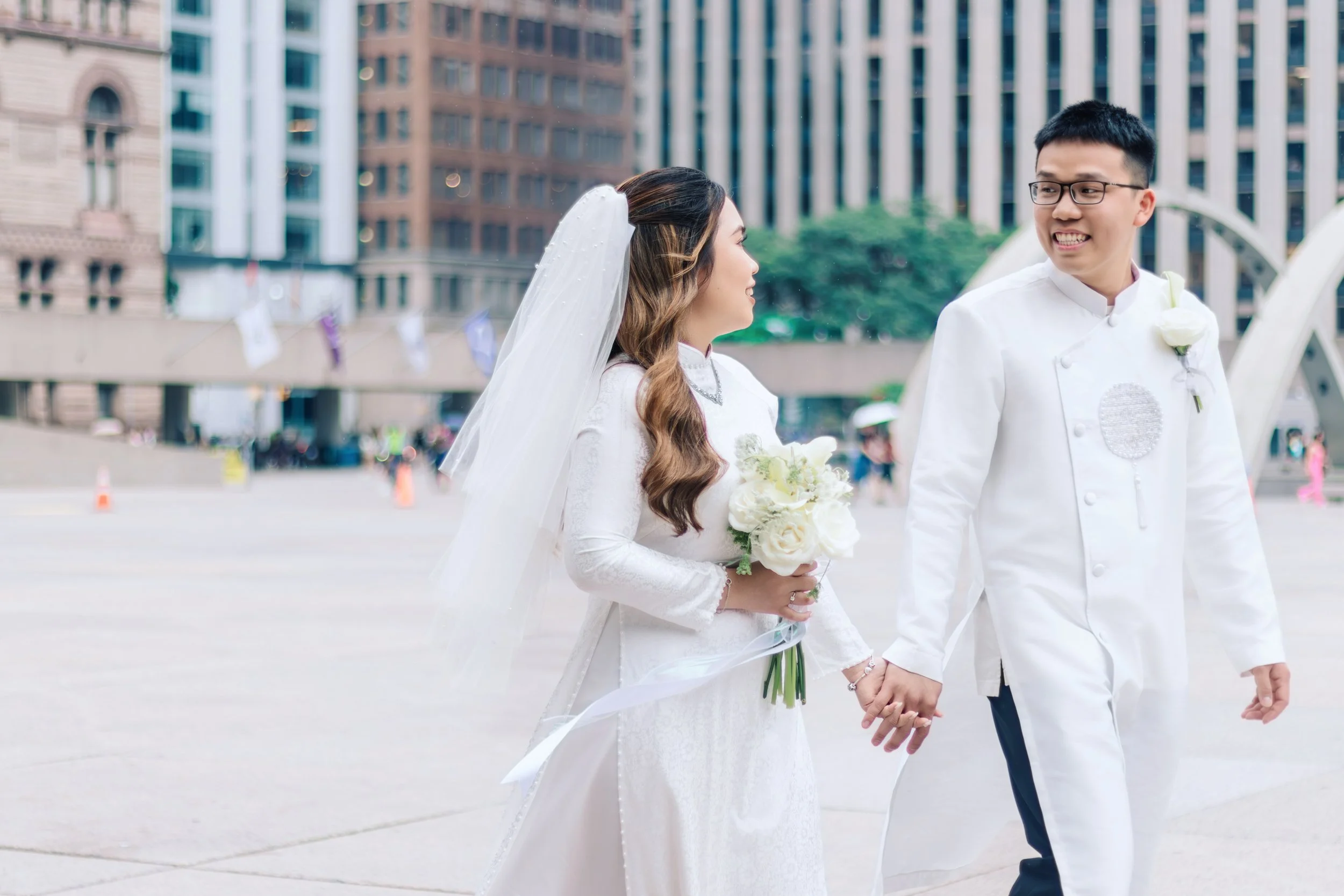 A newlywed couple walking hand in hand in an urban area during their wedding day, with the woman in a white wedding dress holding a bouquet and the man in a white suit with glasses, smiling at each other.