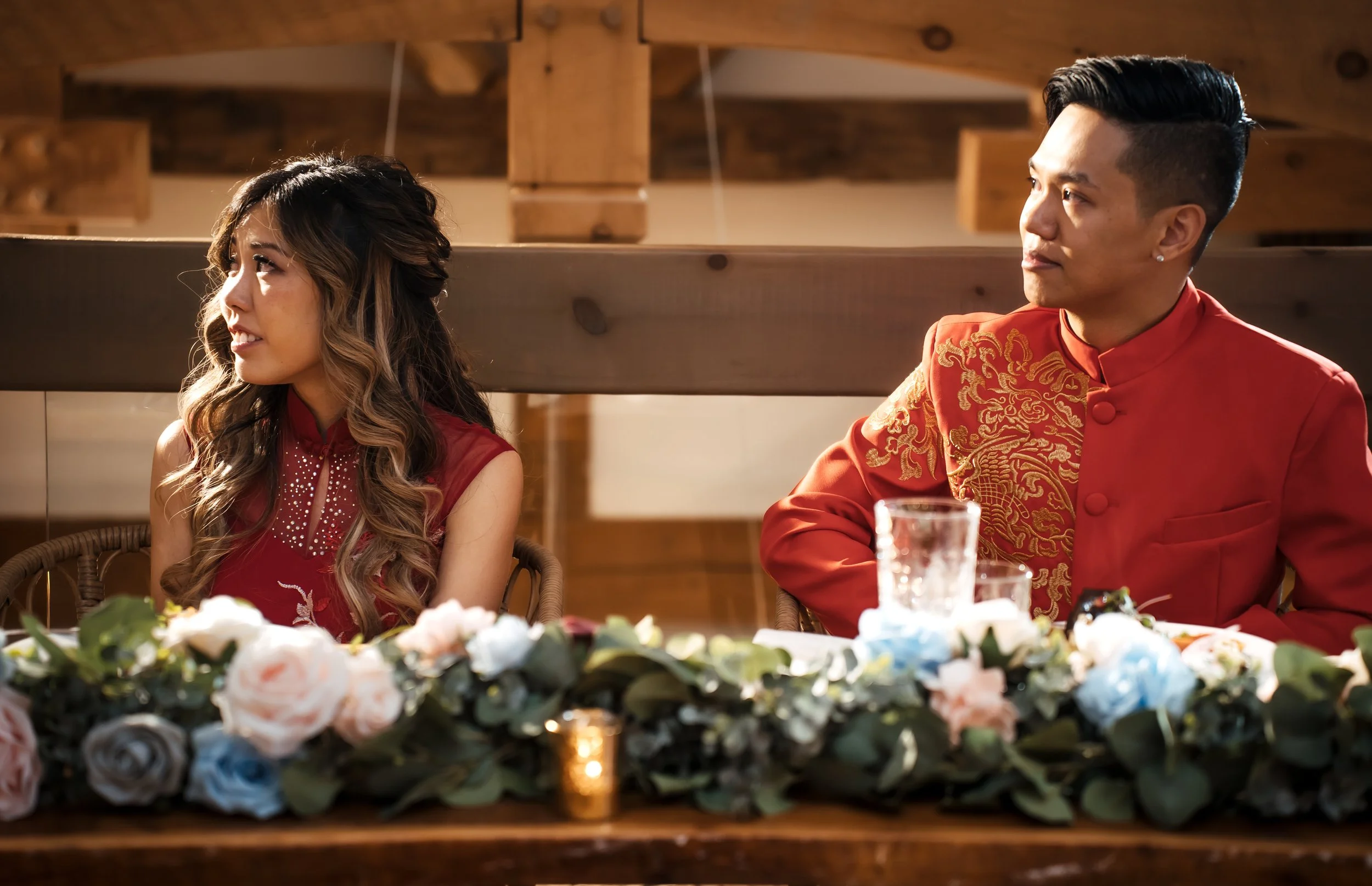 A man and woman sitting at a decorated wedding reception table, looking at each other, with flowers and candle decorations.