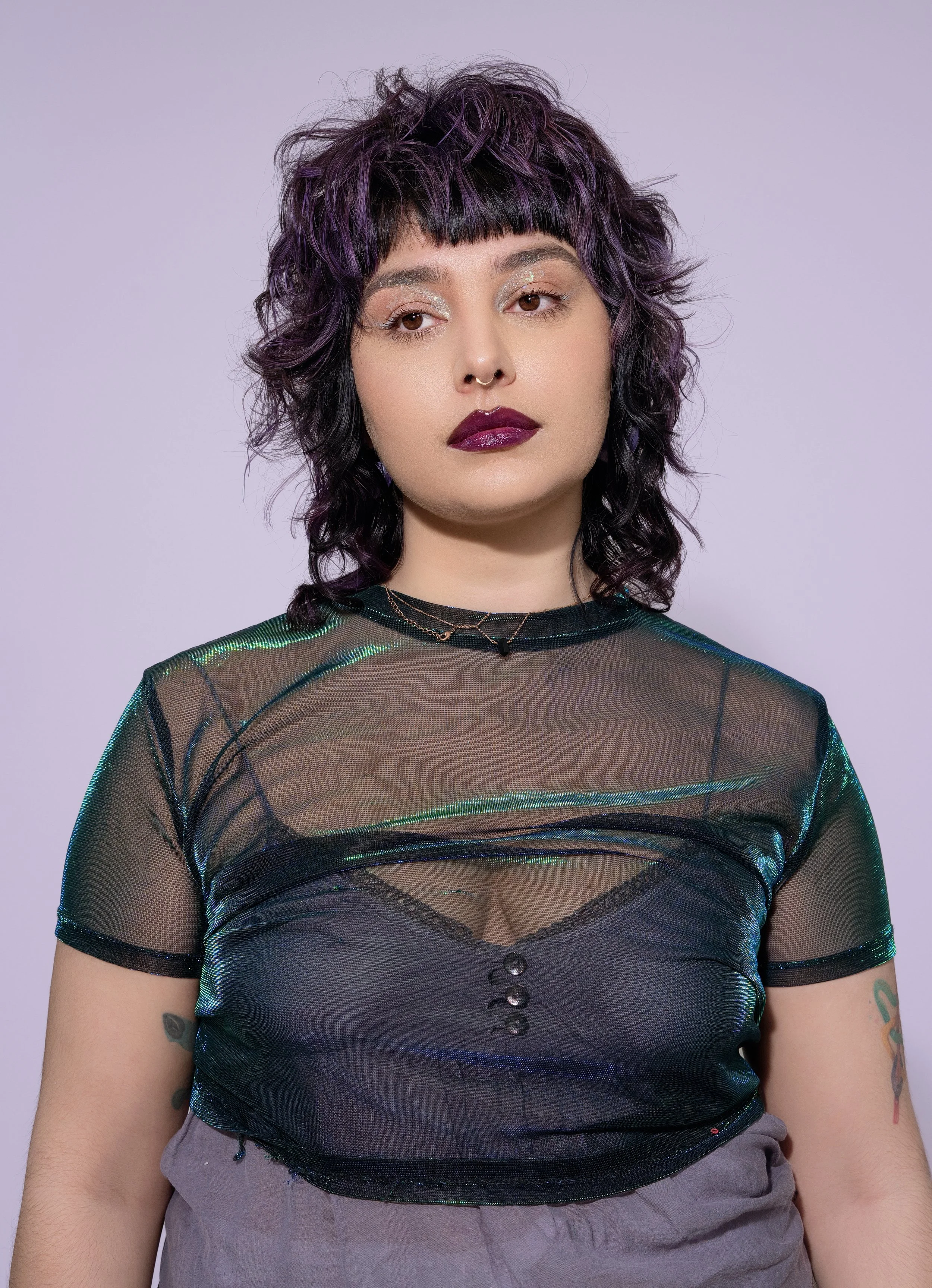 A young woman with curly, shoulder-length purple hair and bangs, wearing a sheer black top over a black bralette, with dark lipstick and eye makeup, standing against a light purple background.