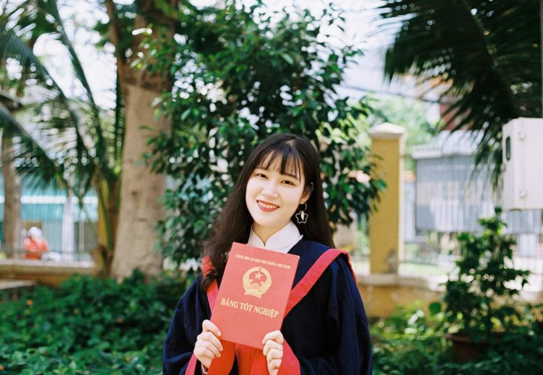A young woman in graduation gown holding a diploma outdoors, smiling.