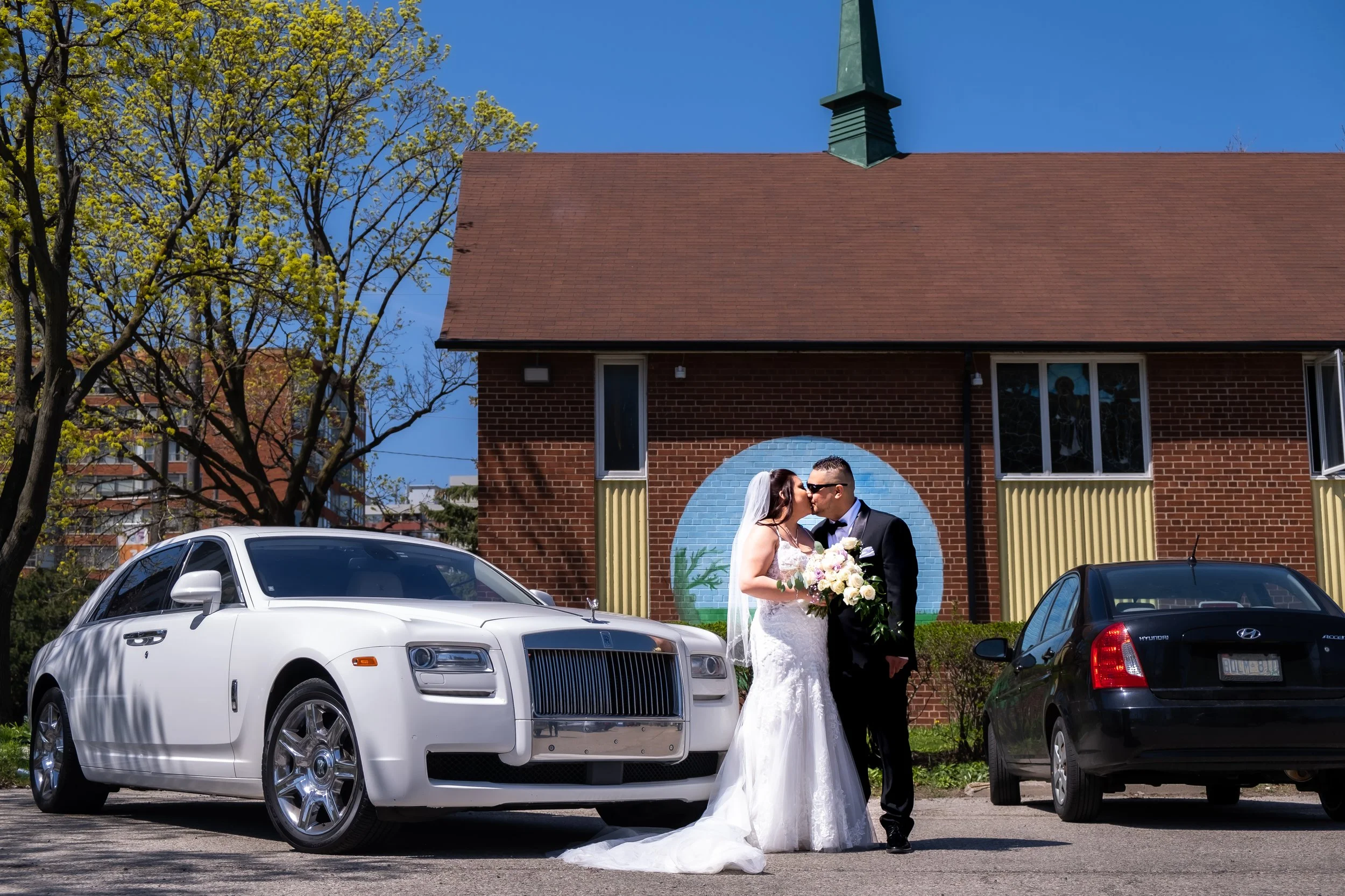 Wedding couple kiss in front of a brick building with a painted mural, flanked by luxury and ordinary cars, and trees with budding leaves in the background.
