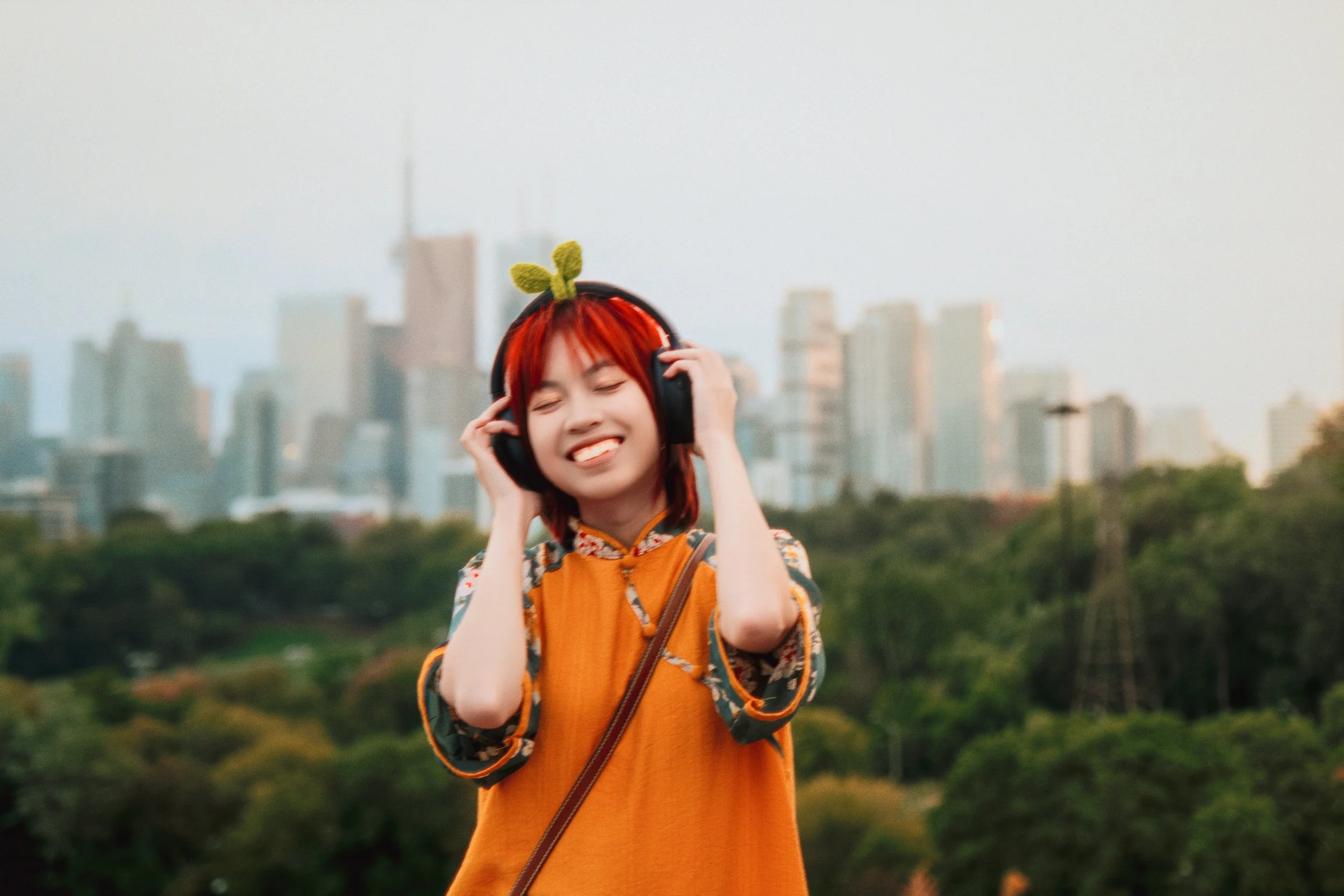 A young woman with red hair and a green hair accessory, smiling and enjoying music with large headphones, standing outdoors with a city skyline and trees in the background.