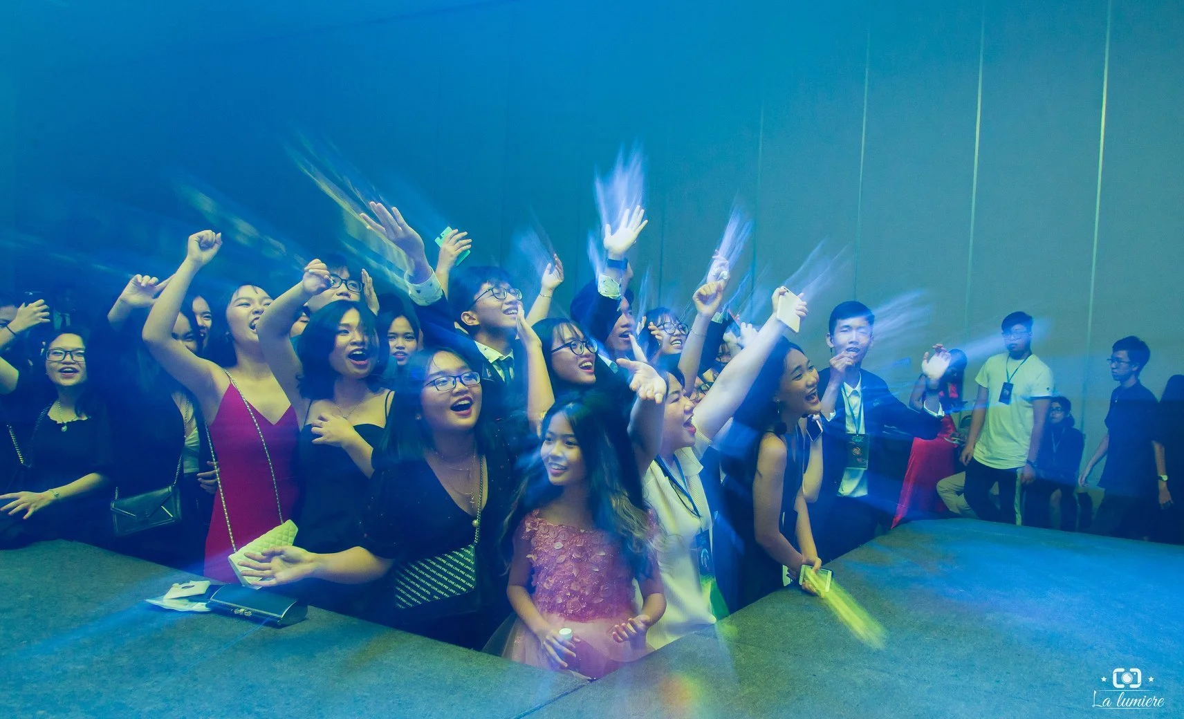 Group of people cheering and celebrating at an indoor event, with some raising their arms and smiling, under blue lighting.