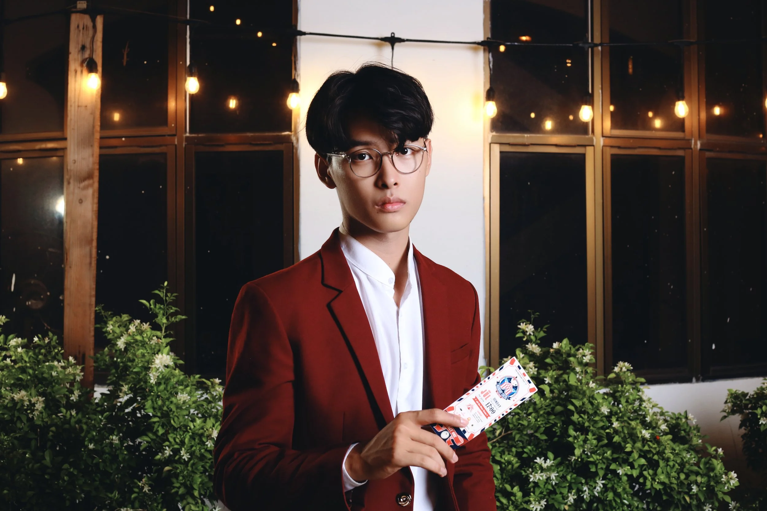 A young man with black hair, glasses, and a red blazer holding event tickets, standing outdoors at night near bushes with white flowers and string lights