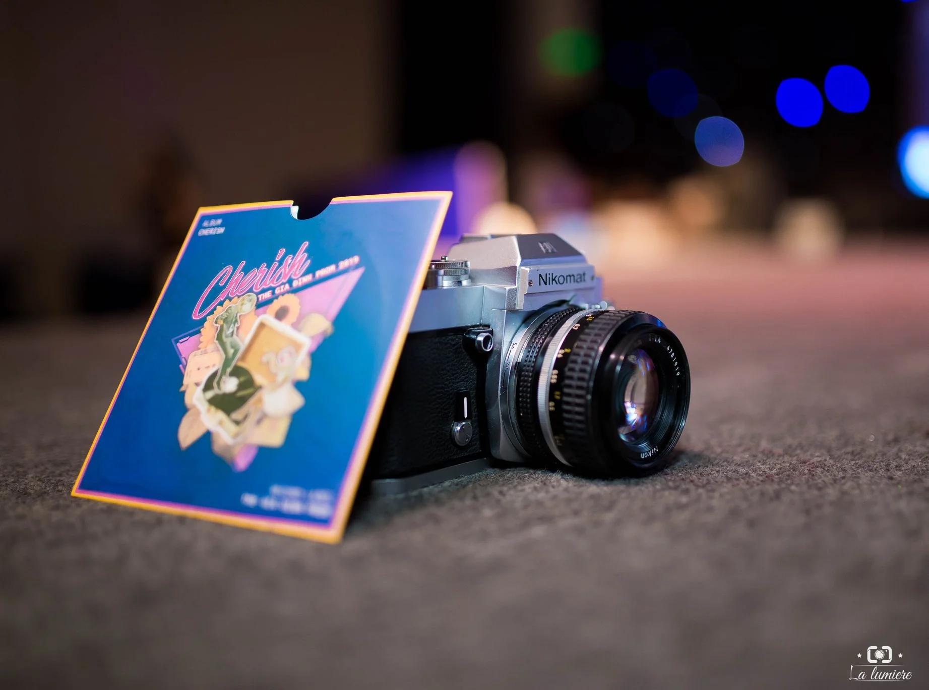 A vintage Nikon camera with a colorful flyer attached to its side, placed on a textured surface with blurred background lights.