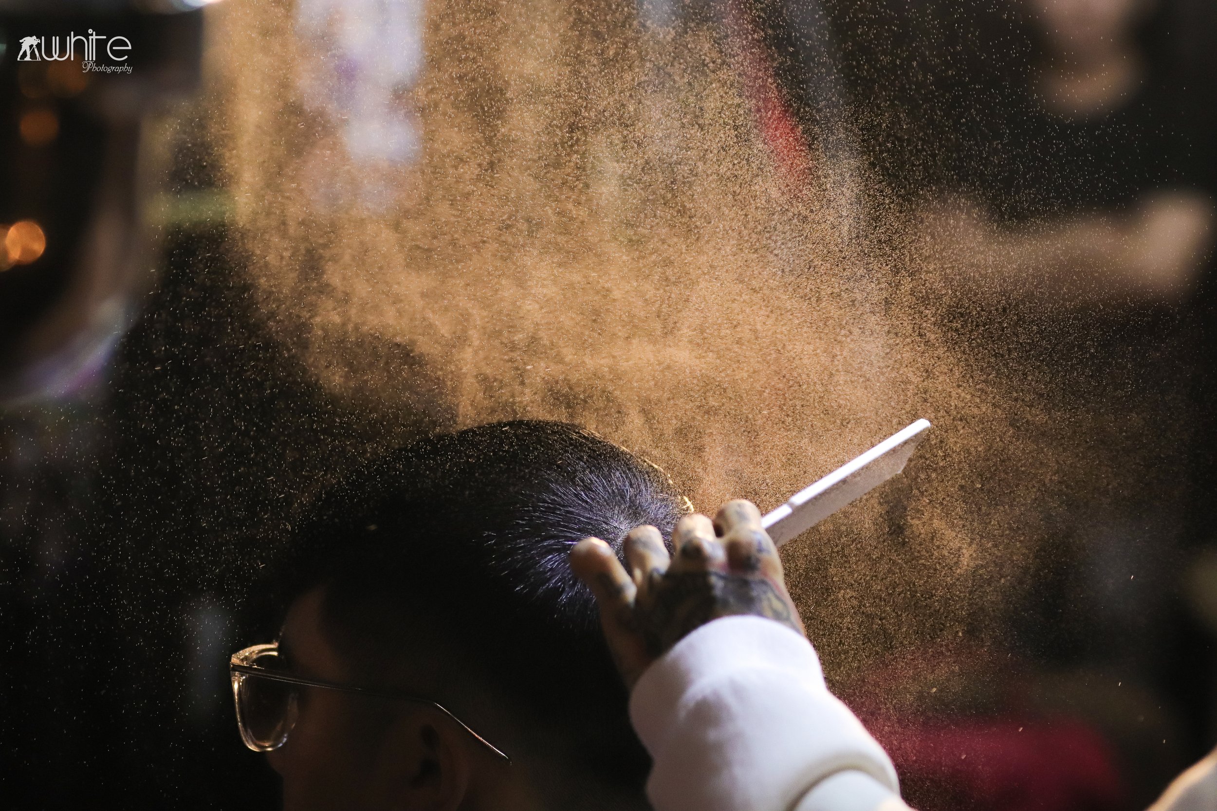 A person with glasses is getting a hair cut, and golden hair clippings are flying through the air.