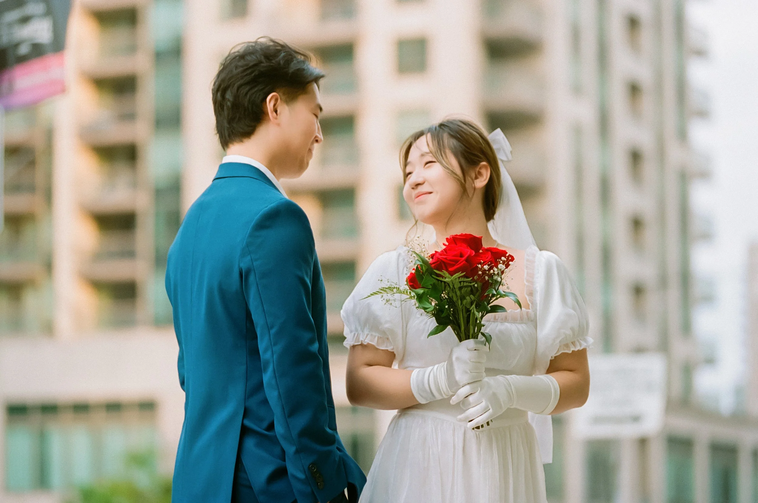 A happy bride and groom on their wedding day, with the bride holding a bouquet of red roses and smiling at her partner.