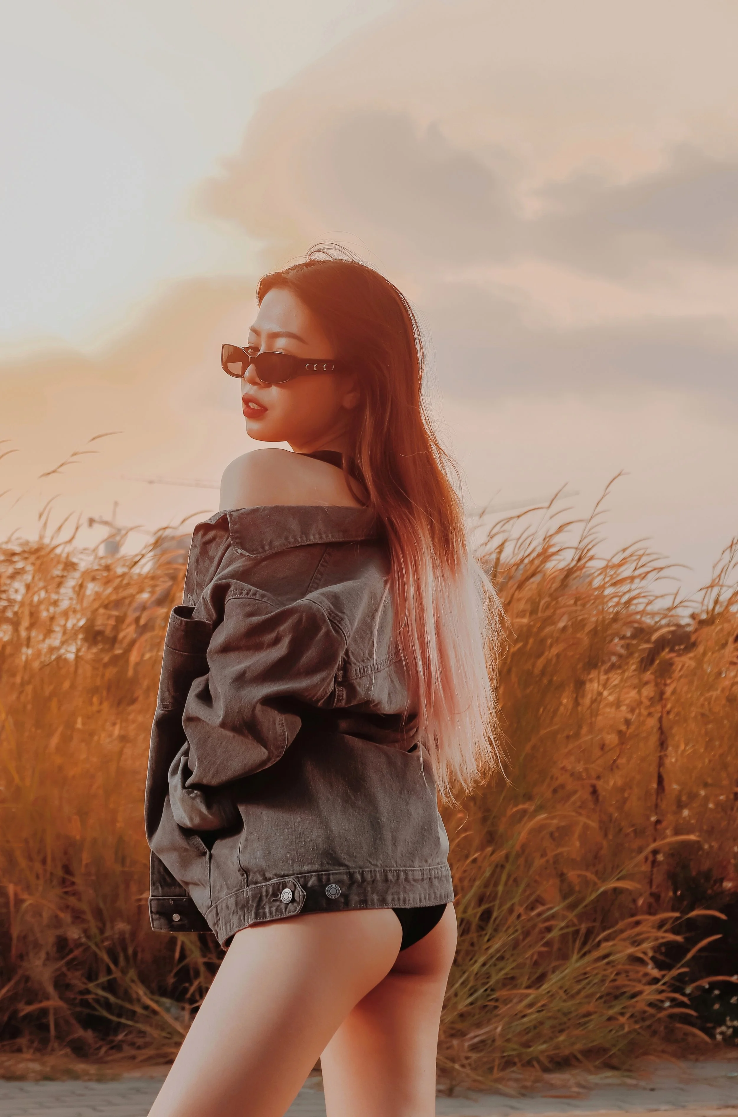 A young woman with long hair, wearing sunglasses and an oversized jacket, standing outdoors in a field with tall grass during sunset, with her back slightly turned and looking over her shoulder.
