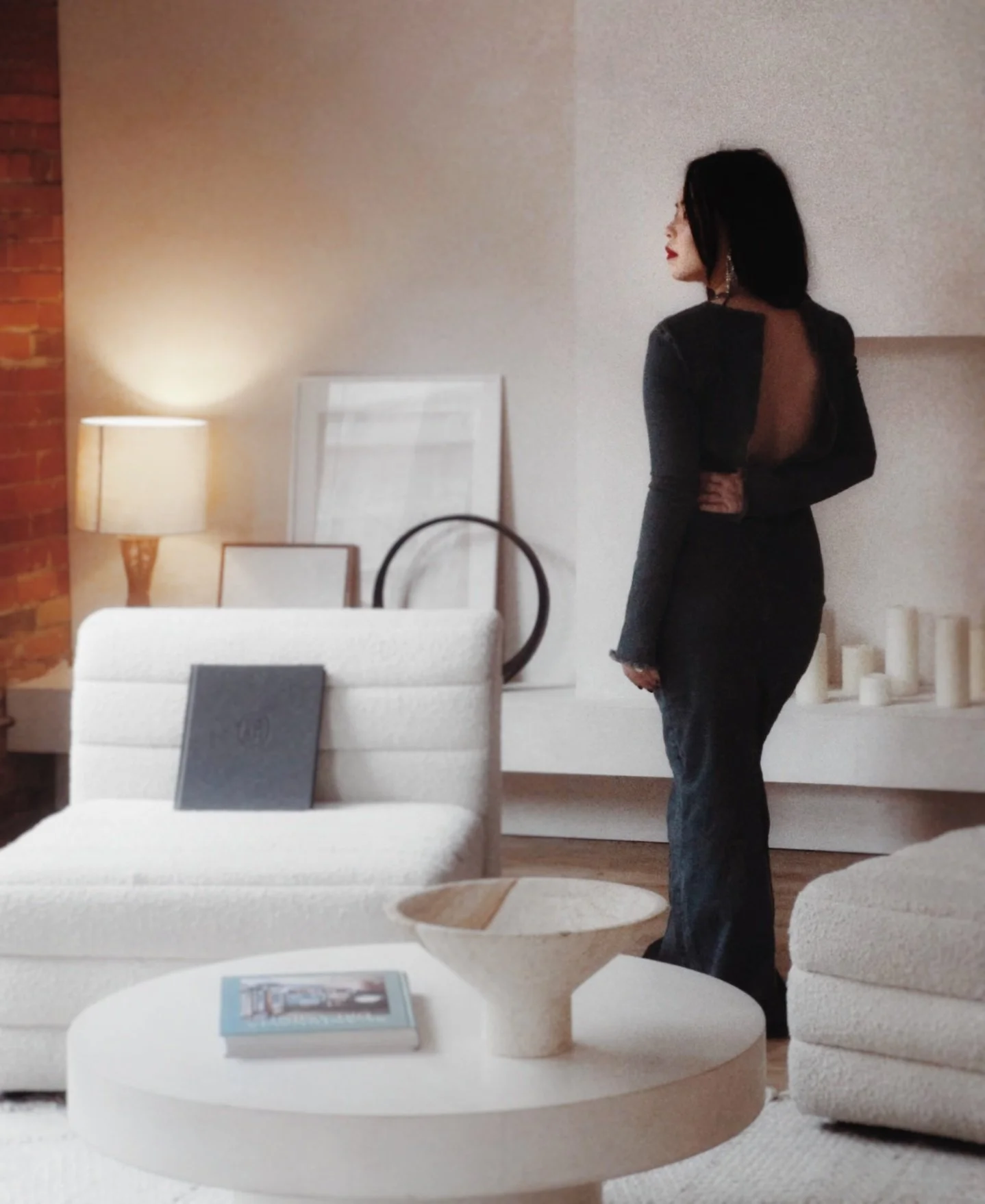 A woman in a black dress stands in a modern living room, facing away from the camera, with white furniture, a lamp, framed artwork, and decorative objects in the background.