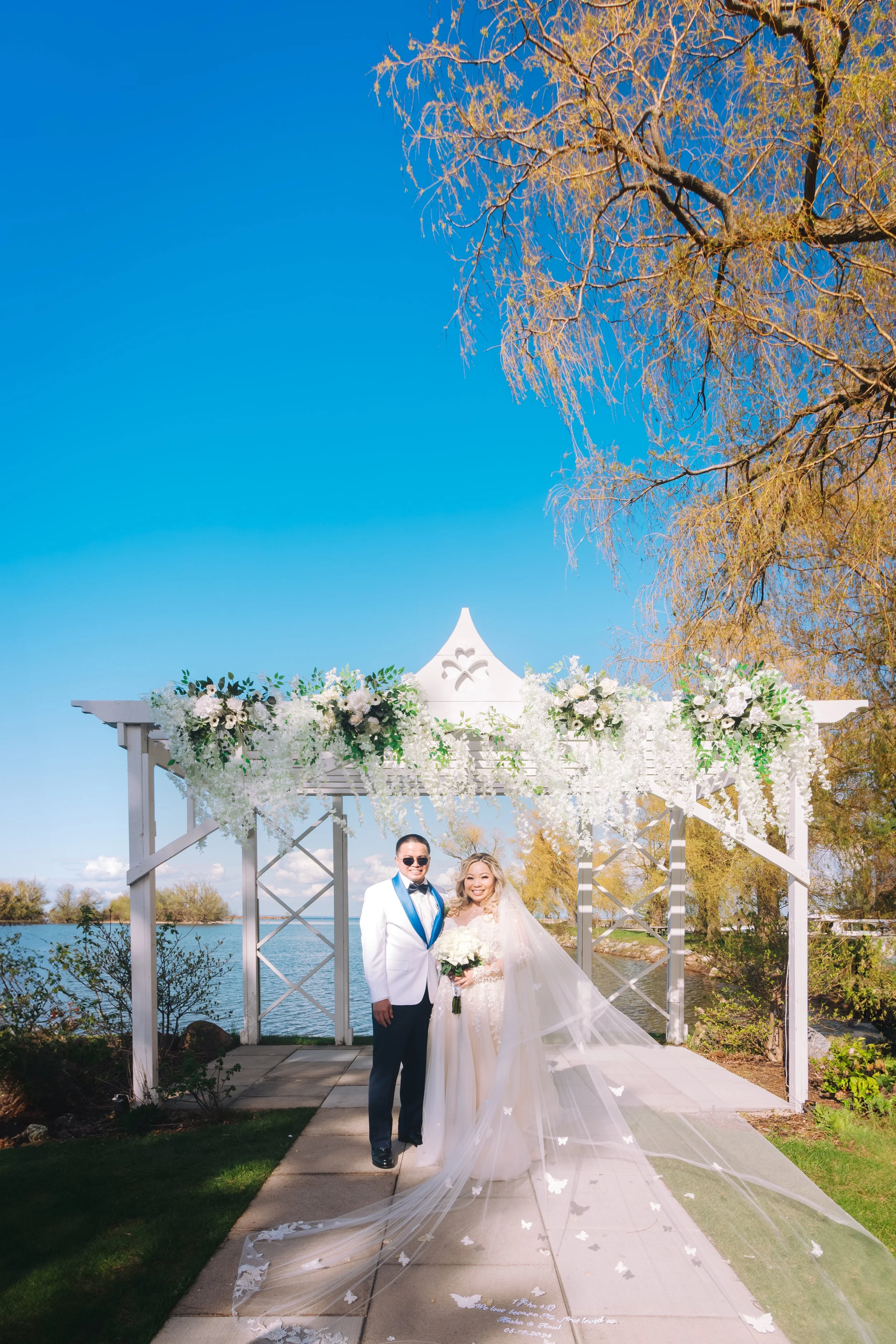 A wedding couple standing outdoors under a white floral arch near a body of water on a sunny day. The groom is dressed in a white tuxedo jacket with blue lapel and black bow tie, and the bride is in a white wedding gown with a long veil holding a bou
