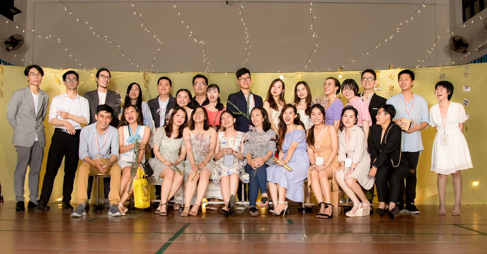 Group of young adults at a celebration or graduation event, posing together on stage with yellow backdrop and fairy lights, some holding flowers and certificates.