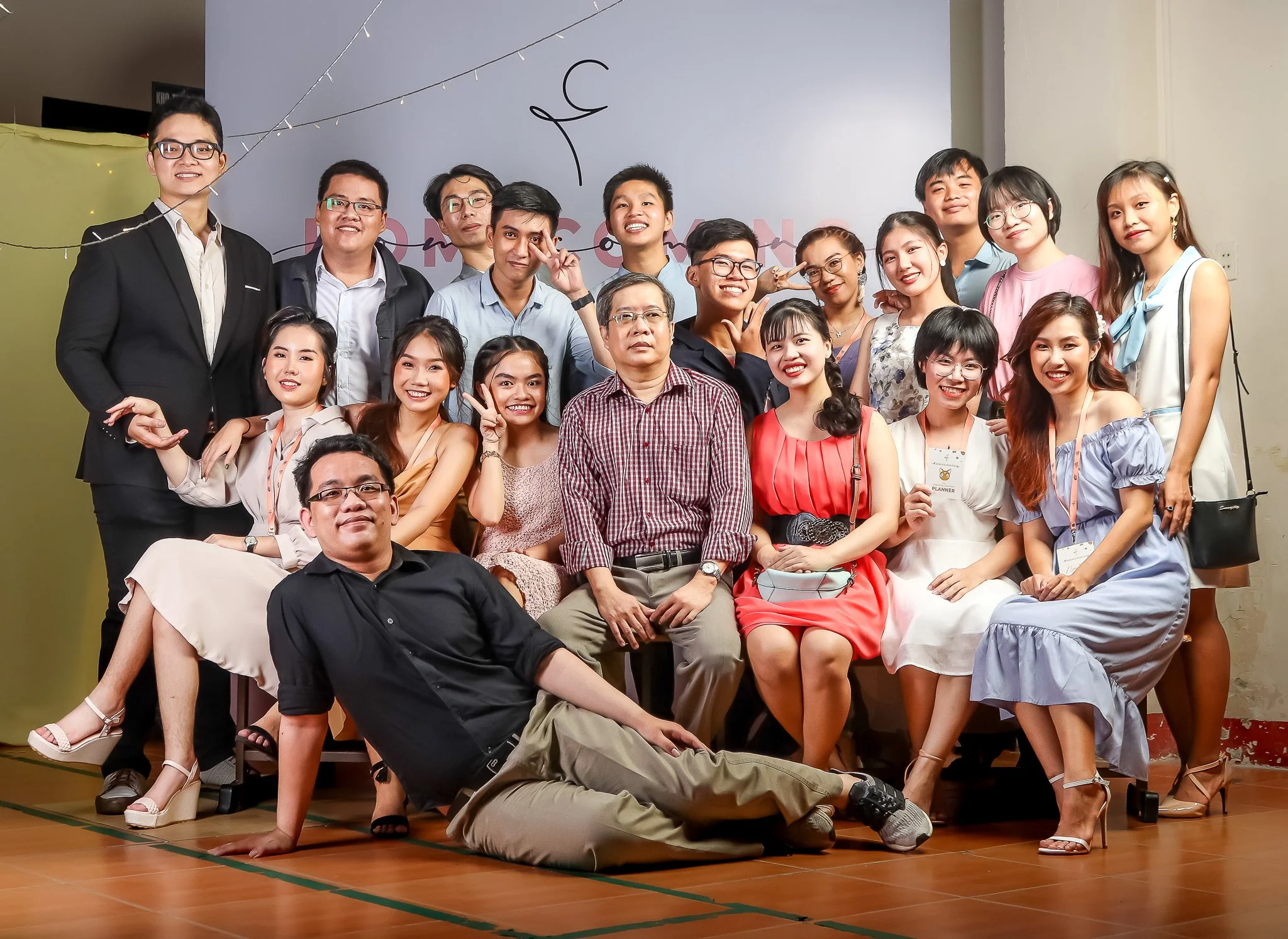 A large group of young adults and one older man are posing for a photo at a celebration event. They are smiling and dressed in semi-formal attire, with some standing and others sitting or lying on the floor. The background features a light-colored wa