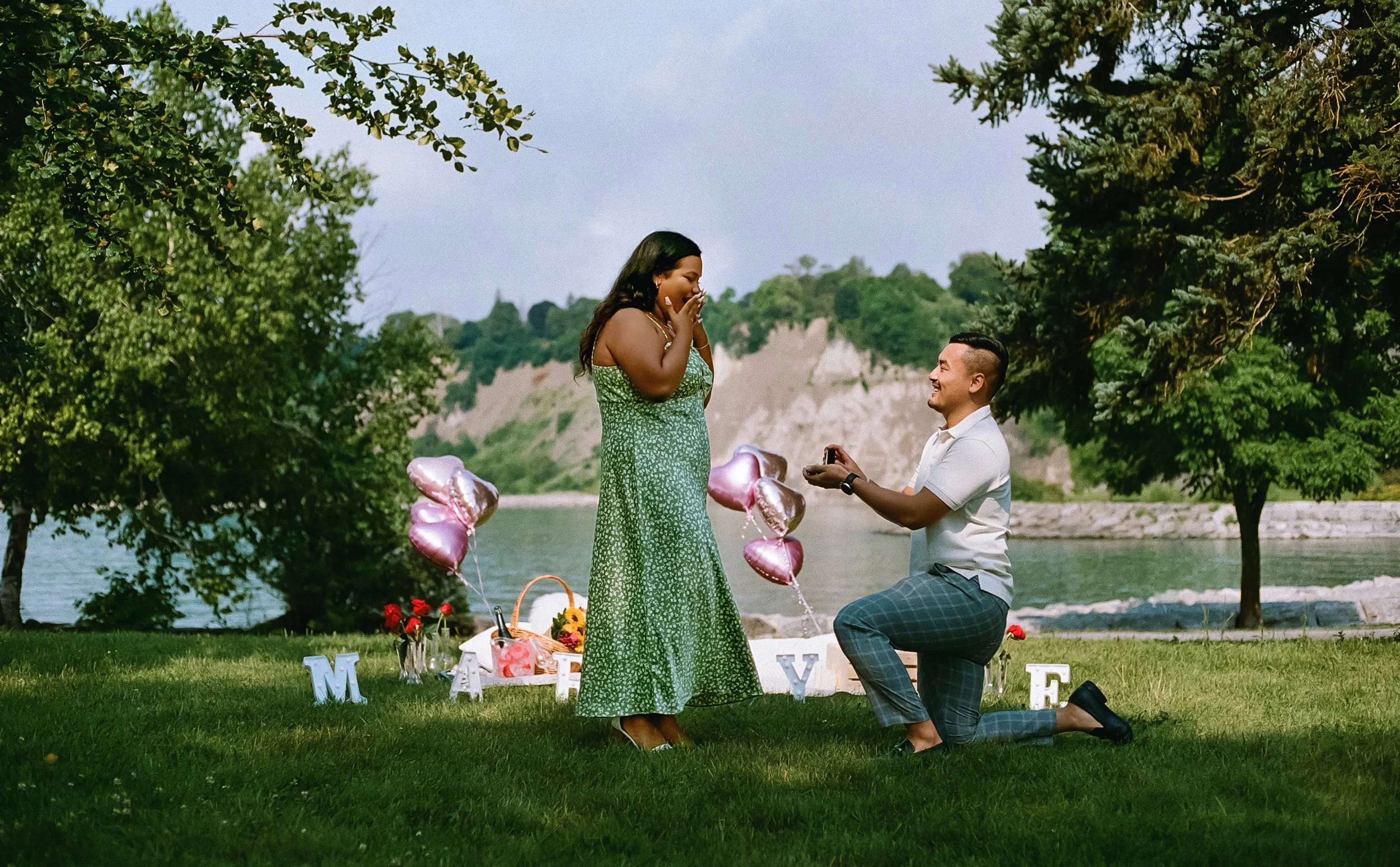 A man proposes marriage to a woman during a romantic outdoor picnic by a lake, with trees, balloons, flowers, and the word 'MARRY' displayed on the grass.