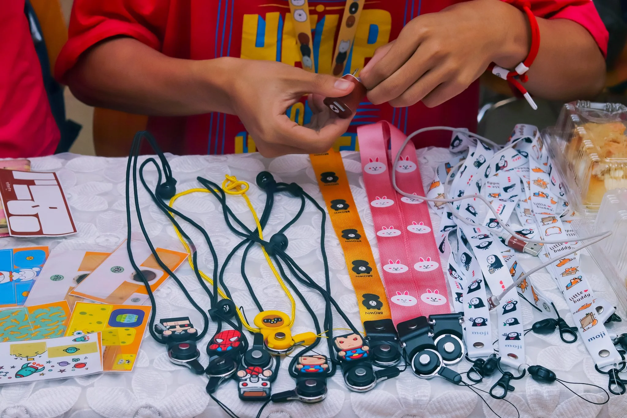 Person handling colorful lanyards and animal-themed or superhero-themed accessories at a display table with various stickers, cables, and snacks.