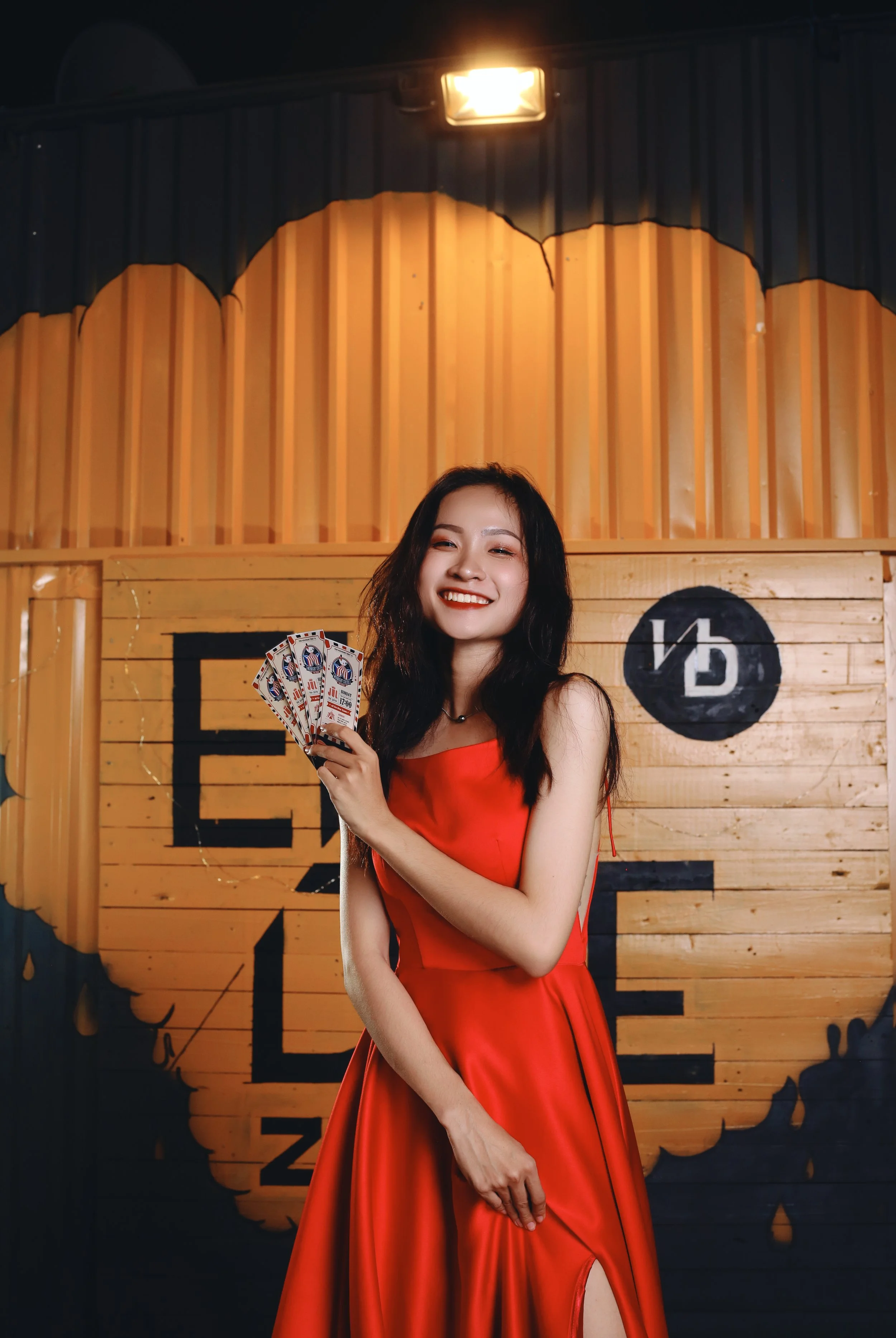 A young woman in a red dress holding playing cards, smiling, in front of a wooden backdrop with black and white graphic design.