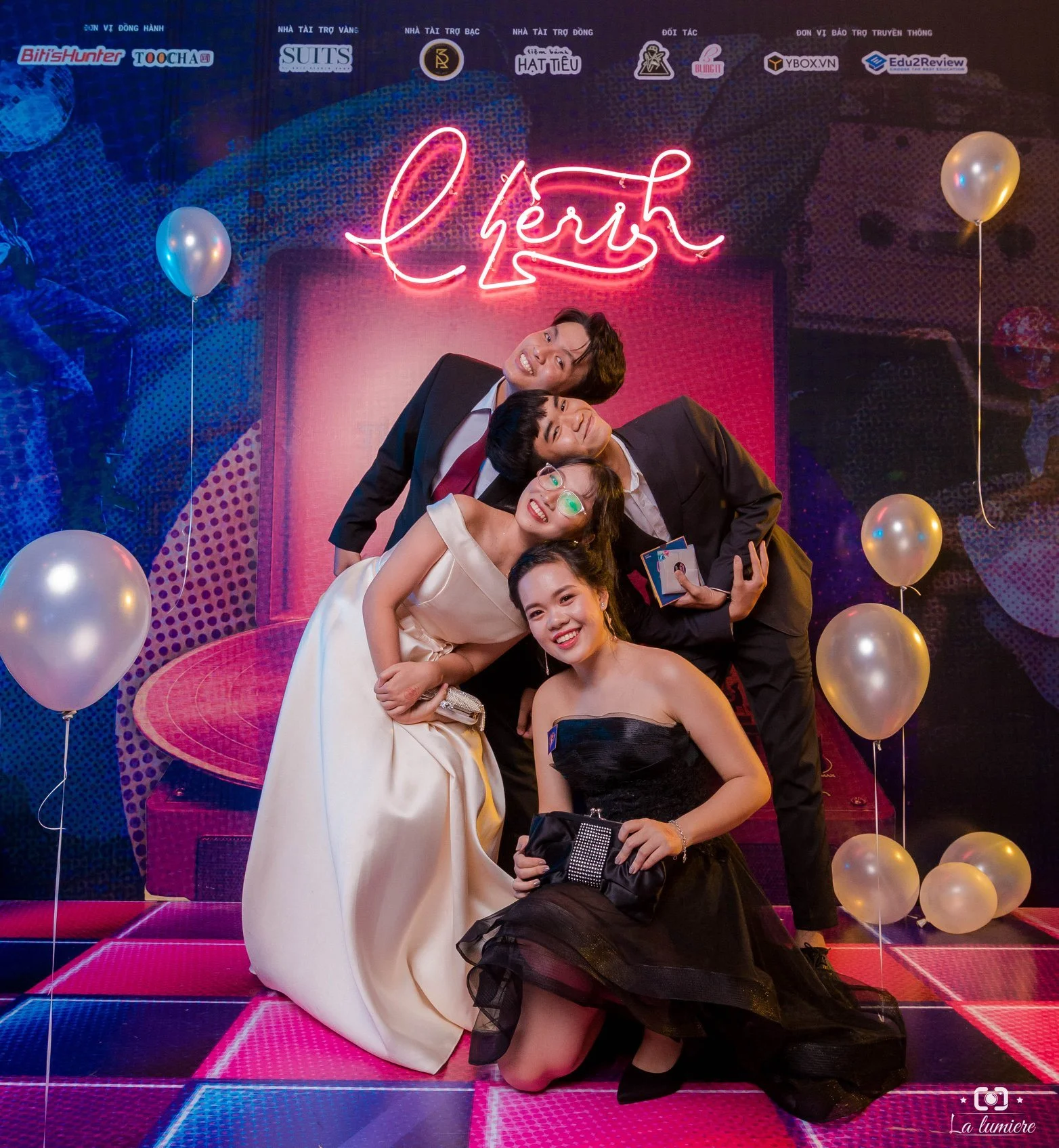 Four people dressed in formal party attire, smiling and posing together in front of a colorful backdrop with balloons and a neon sign reading 'cherish.' The scene appears to be at a celebration or event, with vibrant lighting and a lively atmosphere.