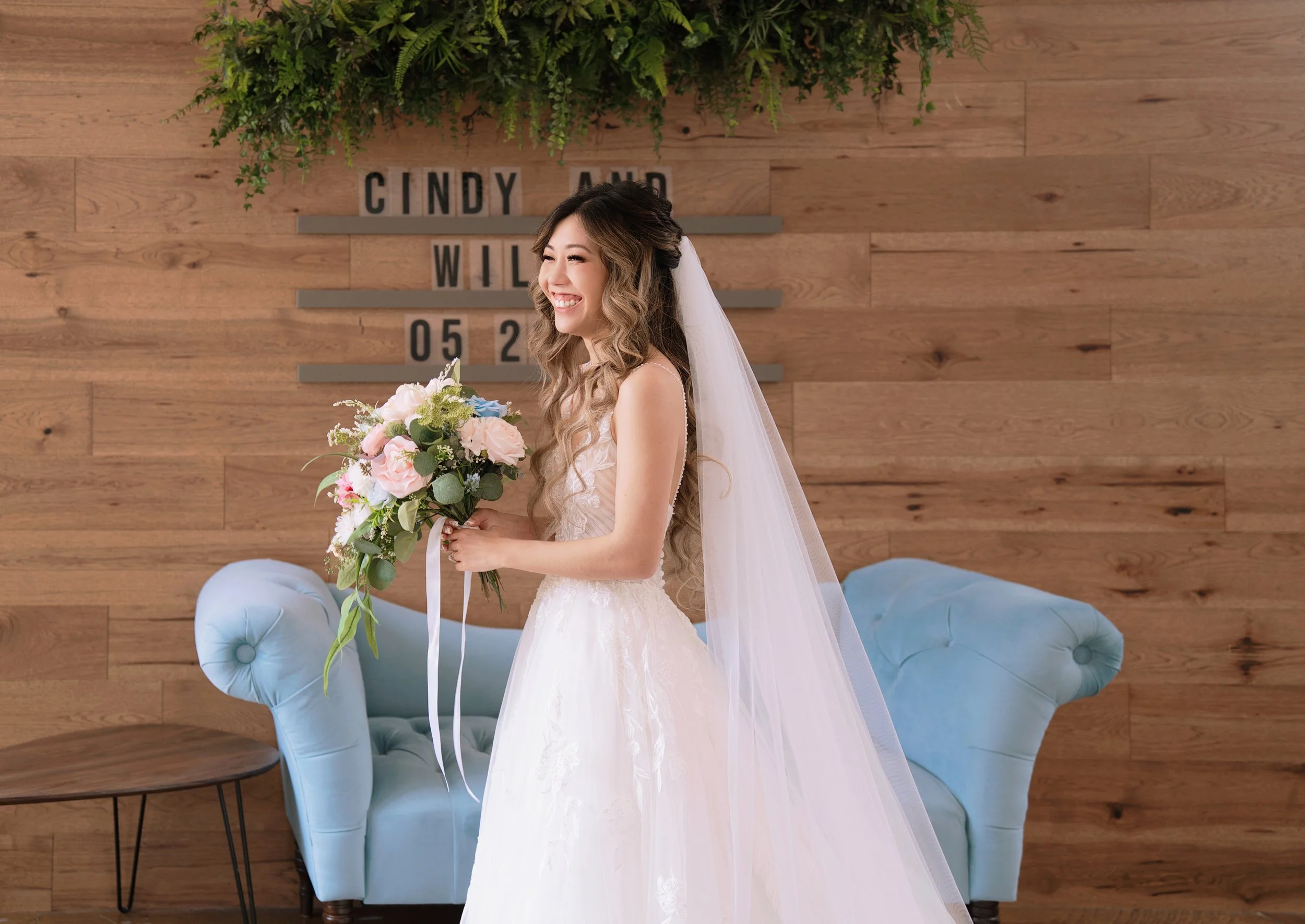 A smiling bride in a white wedding dress holding a bouquet of pink and white flowers, standing in a room with wooden walls and a light blue sofa, with a decorative green plant and a sign behind her.