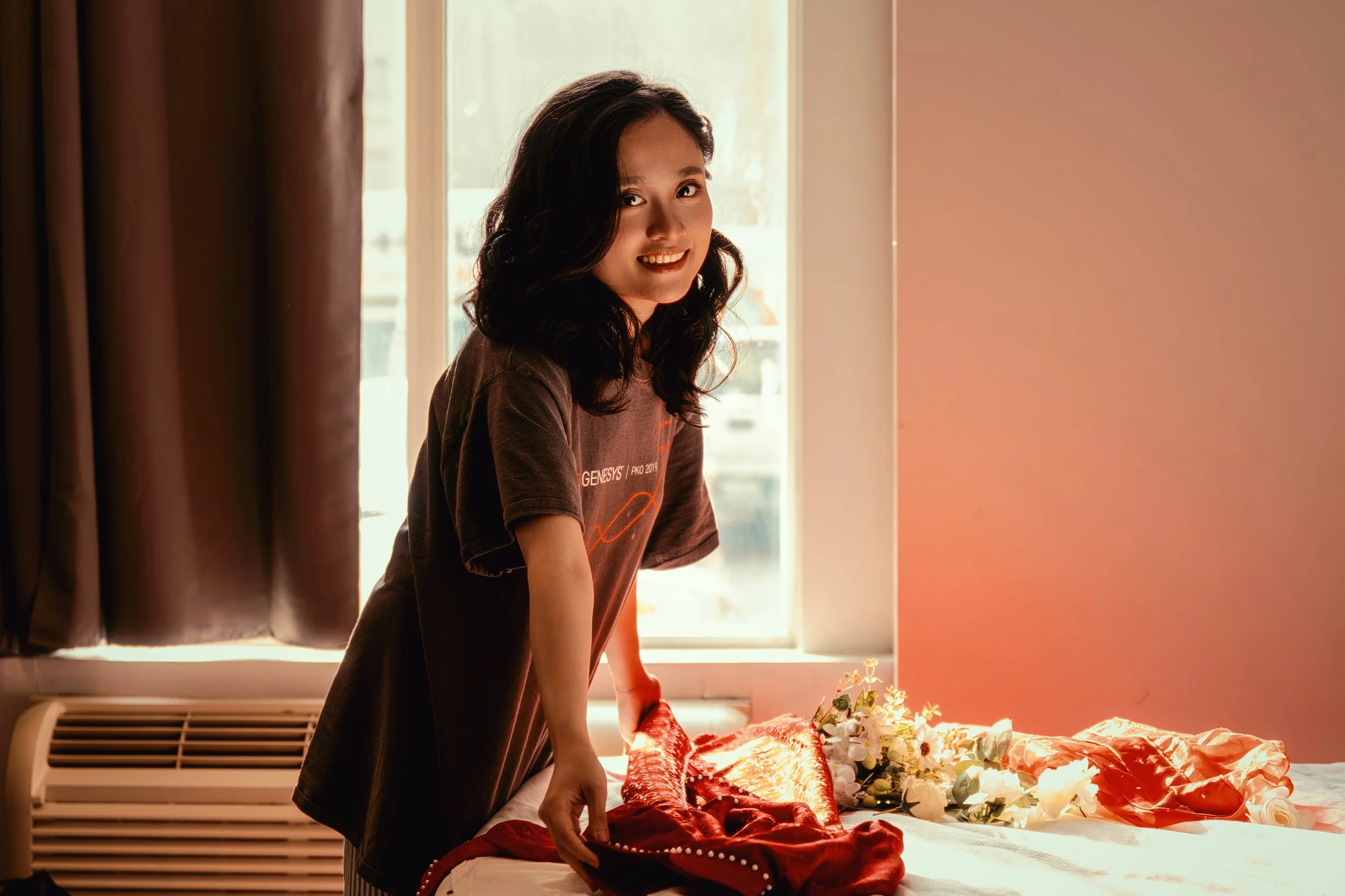 A woman with dark, wavy hair smiling and leaning over a bed decorated with flowers and a red cloth, in a room with a window and beige curtains.