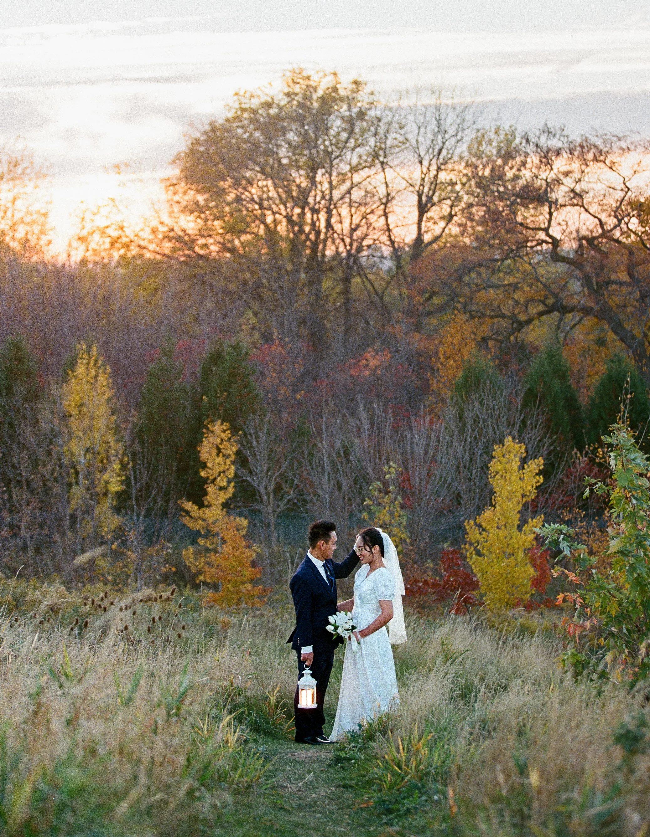 Couple in wedding attire standing on a grassy path in a park with autumn foliage and trees at sunset.