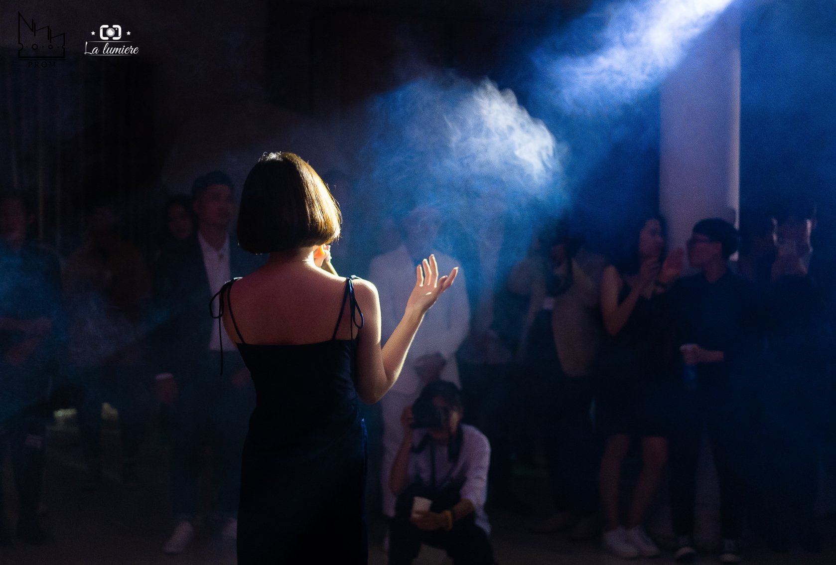 A woman performing on stage at a nightclub, with a microphone in her hand and a black dress, facing a dimly lit audience with some smoke or fog effects.