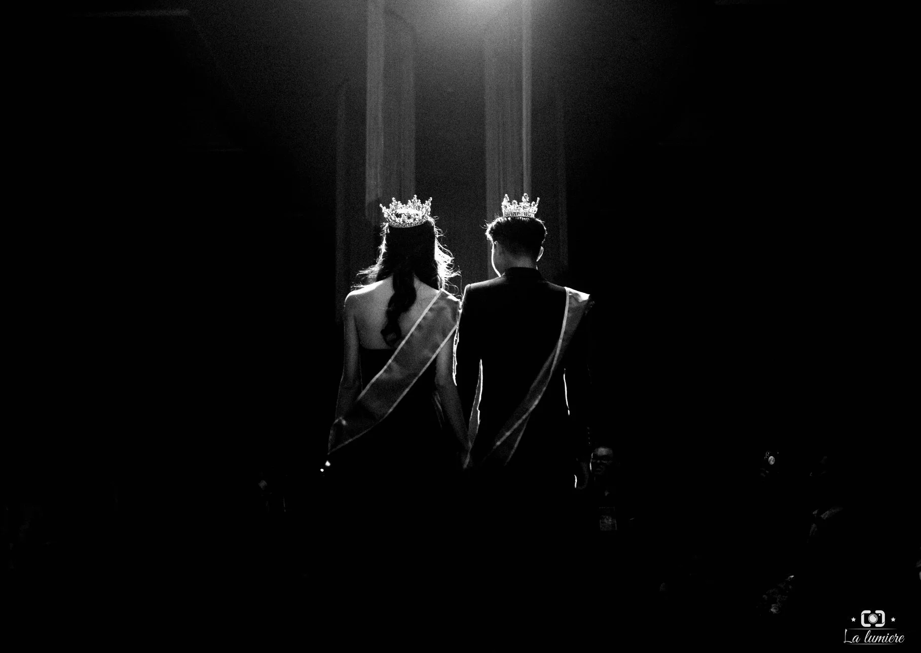 Black and white photo of a couple on stage wearing crowns and sashes, seen from behind, with dramatic lighting highlighting them.