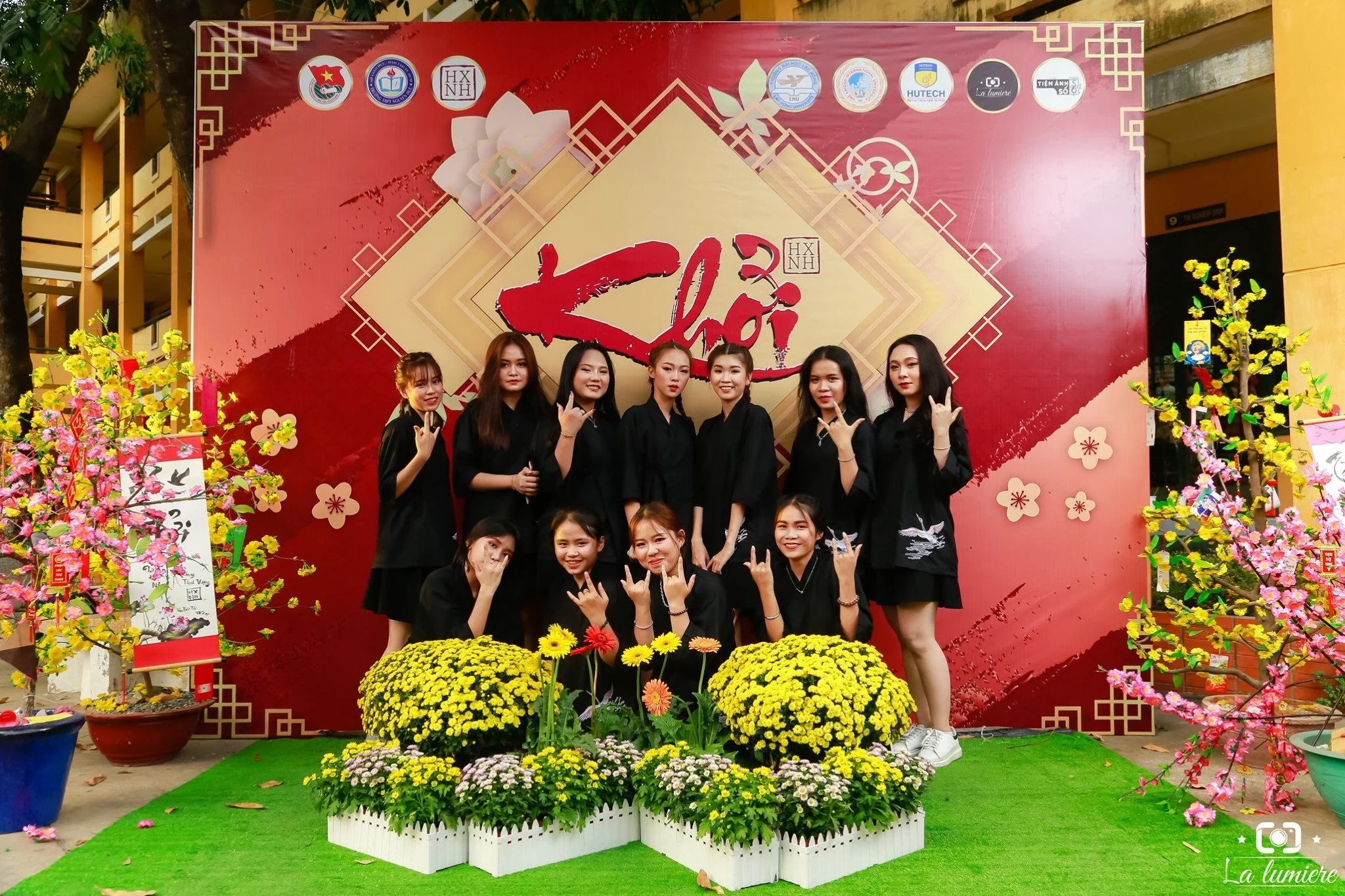 A group of nine young women dressed in black uniforms pose together in front of a red and gold backdrop with floral decorations, during a celebration of the Lunar New Year.