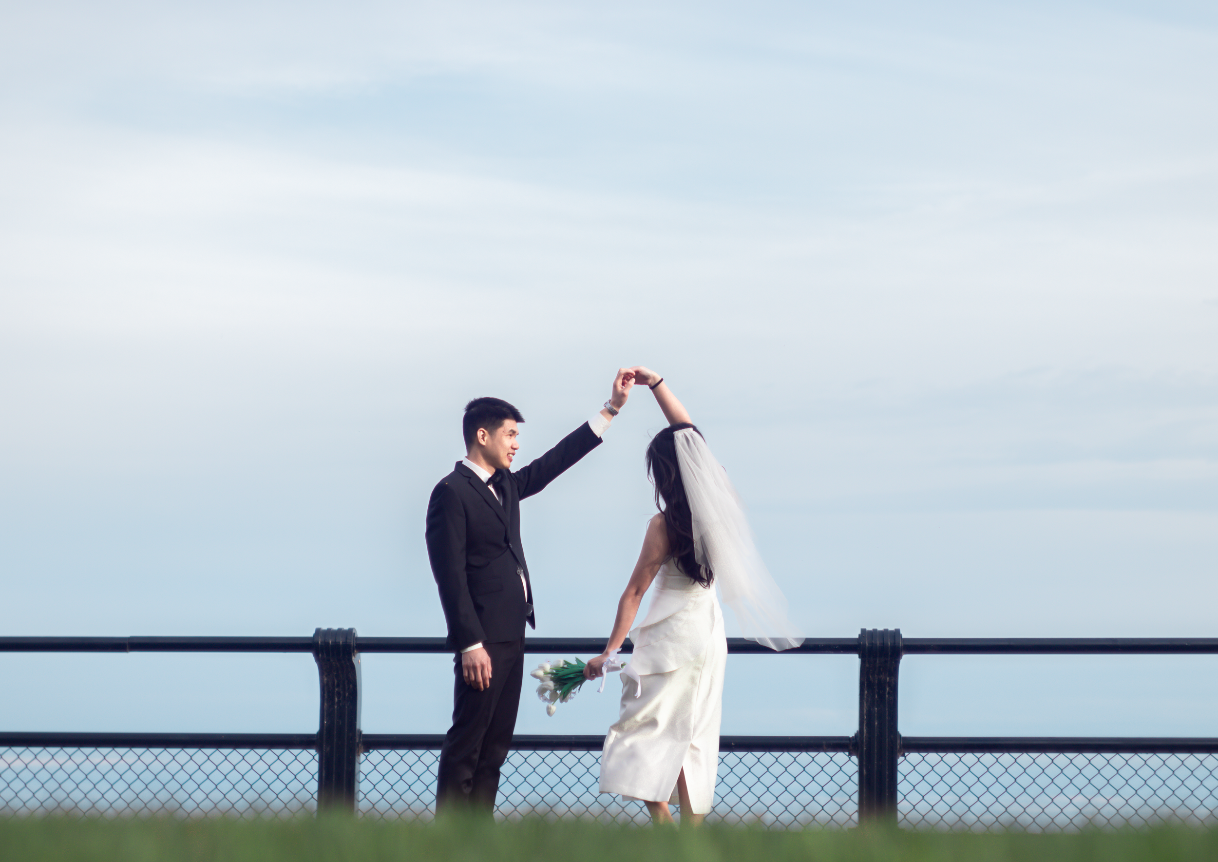 A couple in wedding attire dancing on a platform outdoors, with the man lifting the woman's arm. The woman is holding a bouquet of white flowers, and the background features a partly cloudy sky.
