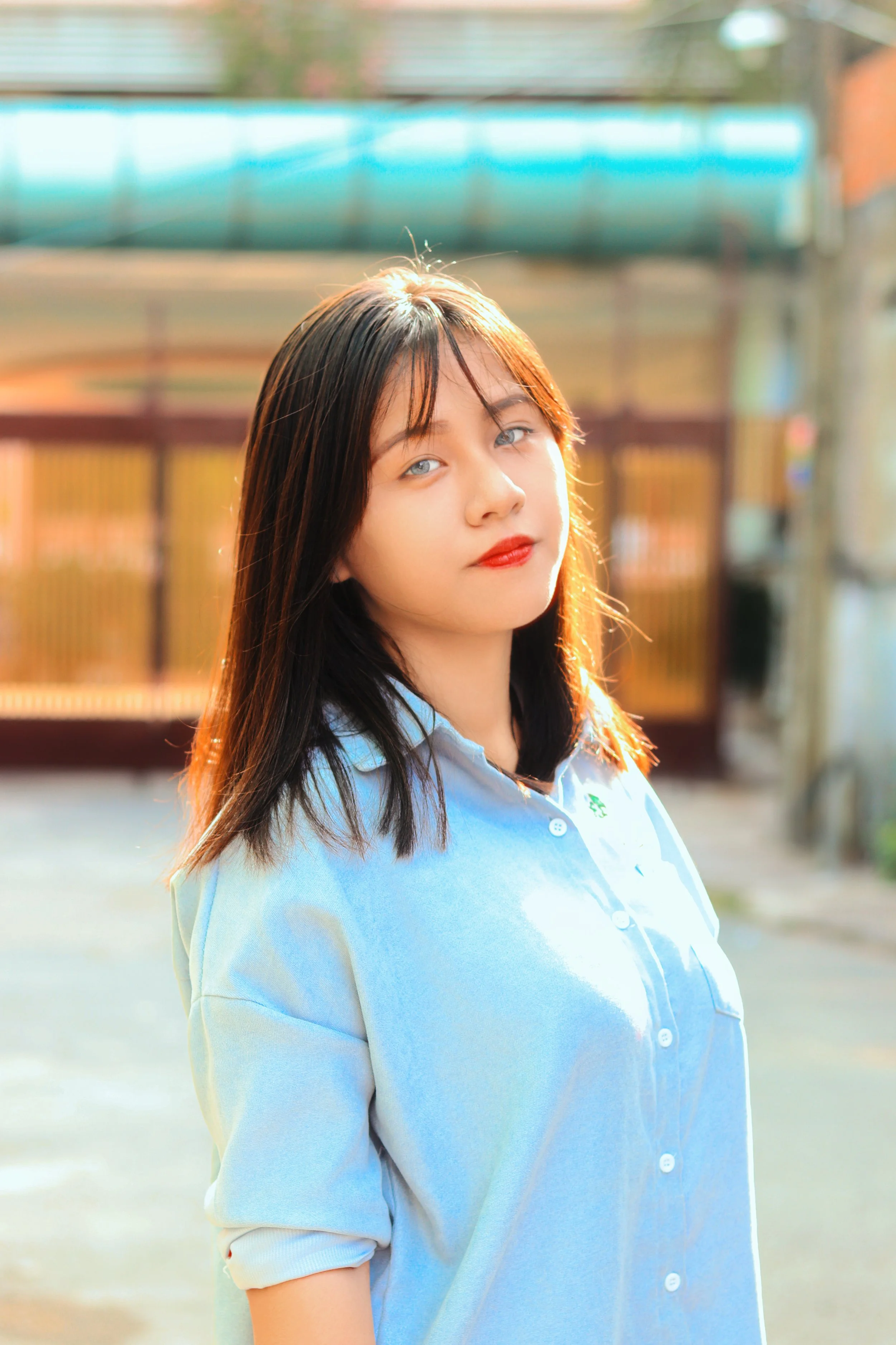 A young woman with dark hair and light blue eyes wearing a light blue button-up shirt standing outdoors in sunlight, with a blurred background of a fence and trees.