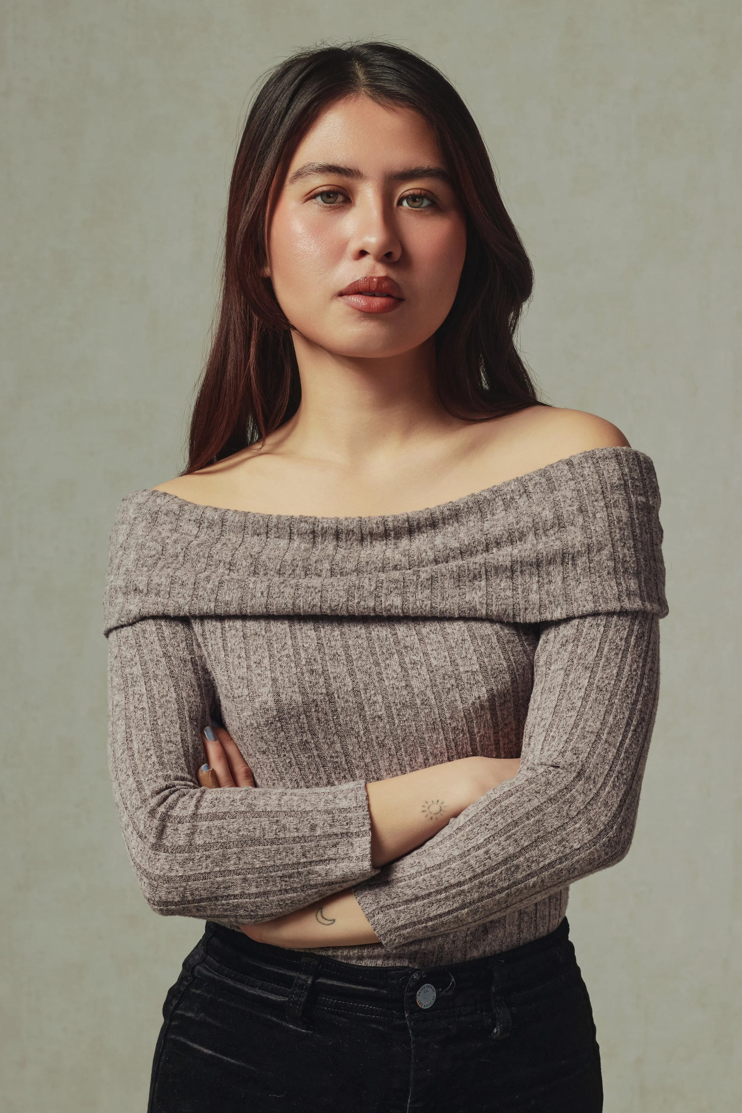 A woman with long dark hair and light skin, wearing a gray off-shoulder sweater and black pants, crossing her arms, standing against a textured beige background.