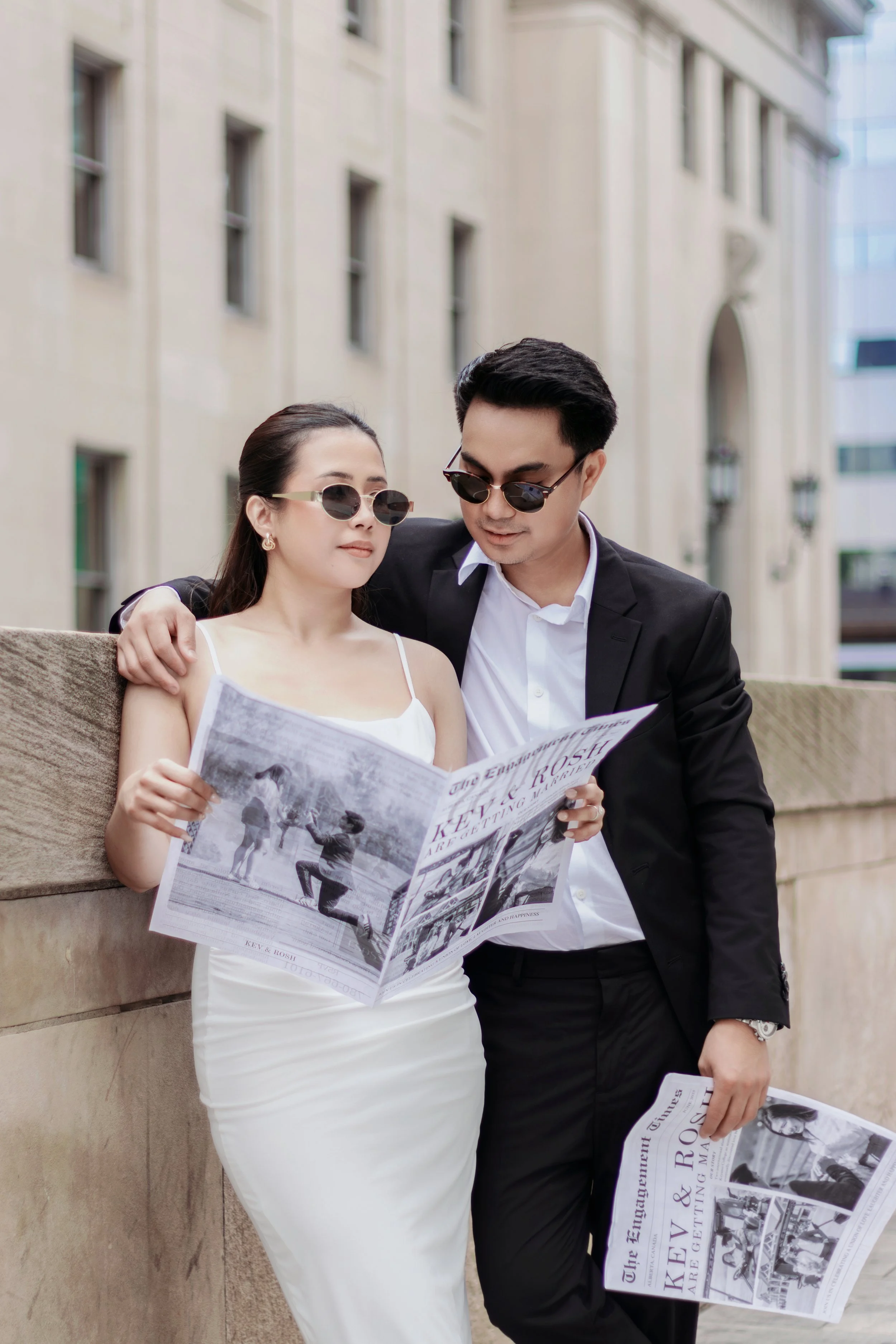 A stylish couple wearing sunglasses standing outdoors, reading newspapers, with the woman leaning against a stone ledge and the man with his arm around her shoulders.