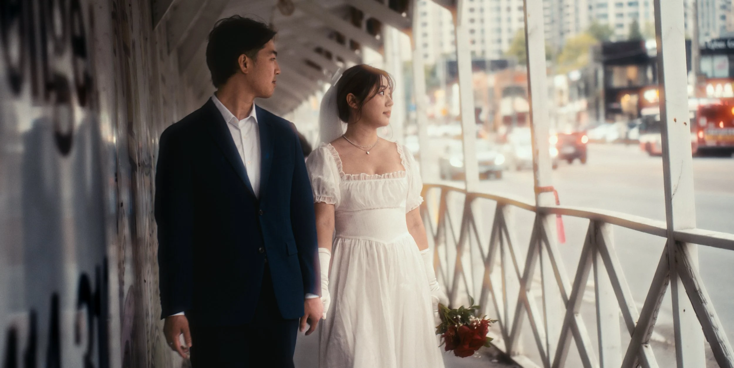 A bride in a white wedding dress holding a bouquet and a groom in a dark suit standing in an outdoor walkway by a city street.