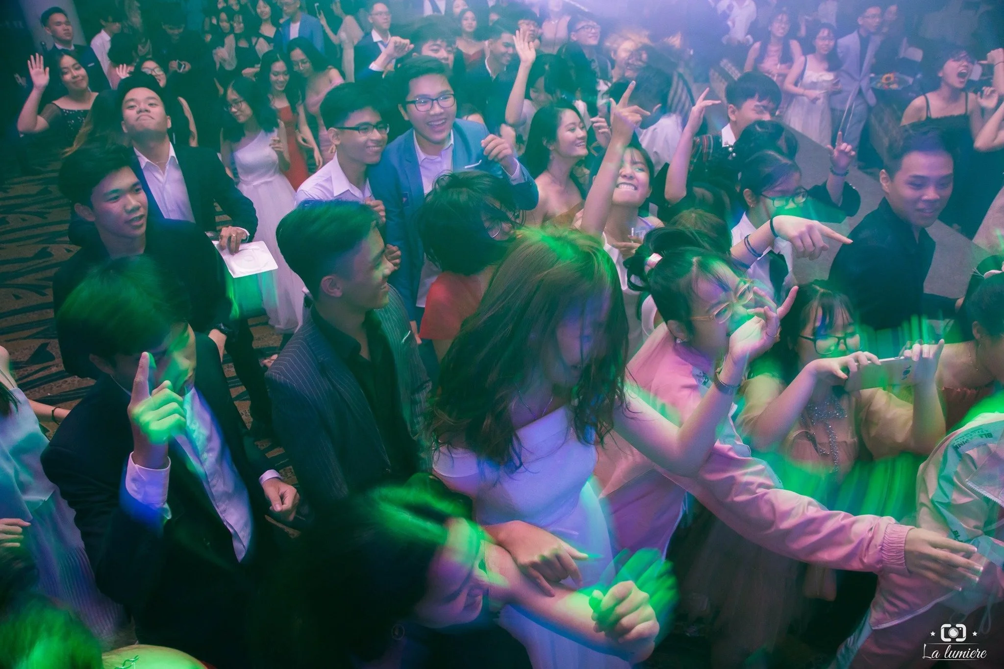 A large group of young people at a lively event or concert, some dancing, singing, and taking photos, with colorful lighting effects.