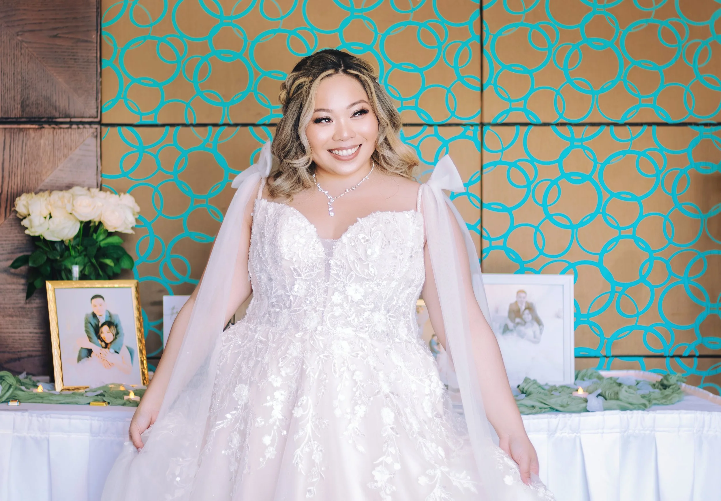 A woman in a white wedding dress with lace details, smiling, wearing a necklace, in front of a modern decorative background, with framed photos and flowers on a table behind her.