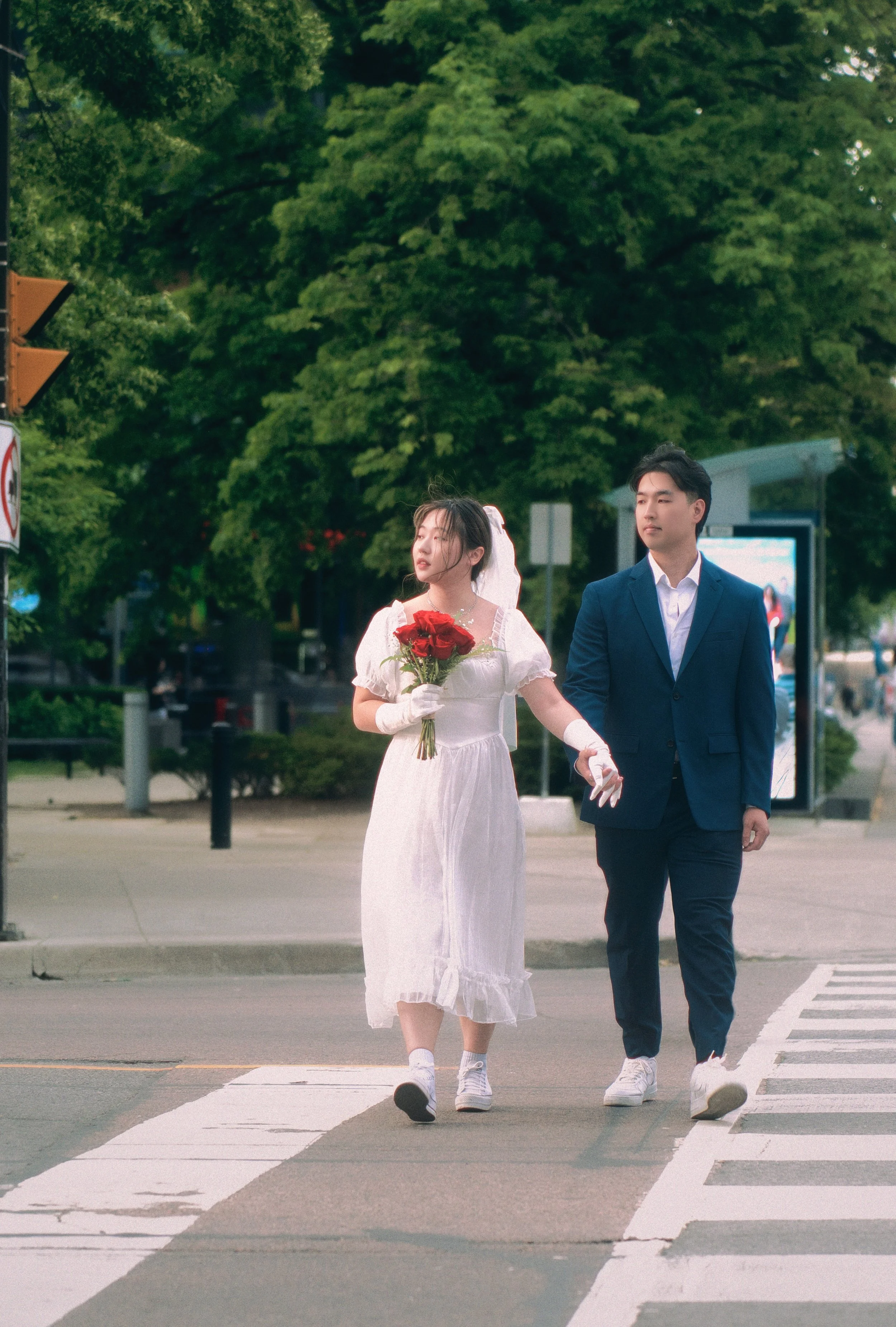 A couple, possibly newlyweds, walking across a crosswalk on a city street. The woman is wearing a white dress and gloves, holding a bouquet of red roses, and is holding hands with the man. The man is dressed in a blue suit and white shirt. There are 