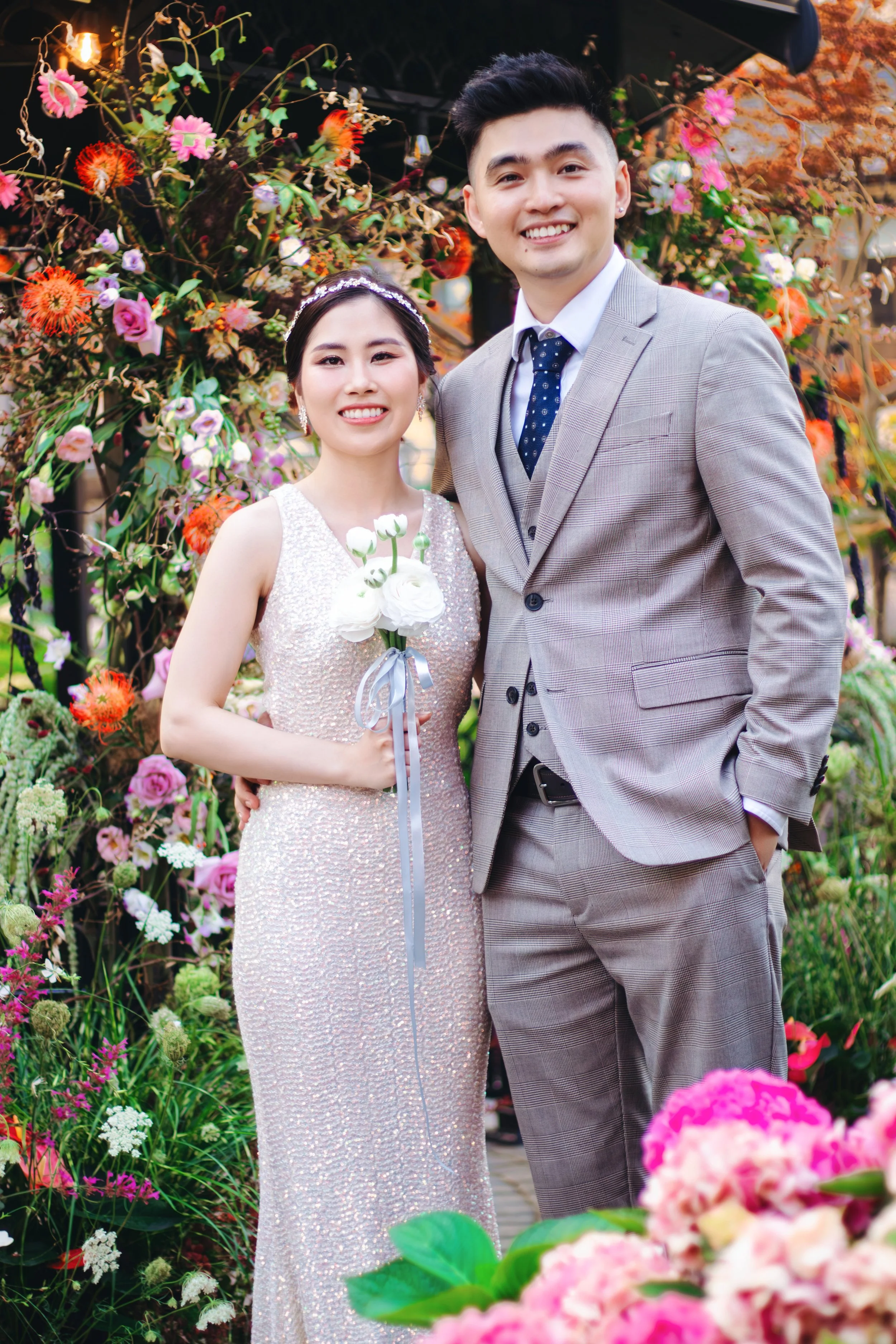 A bride and groom smiling at the camera outdoors, surrounded by colorful flowers and greenery during a wedding celebration.